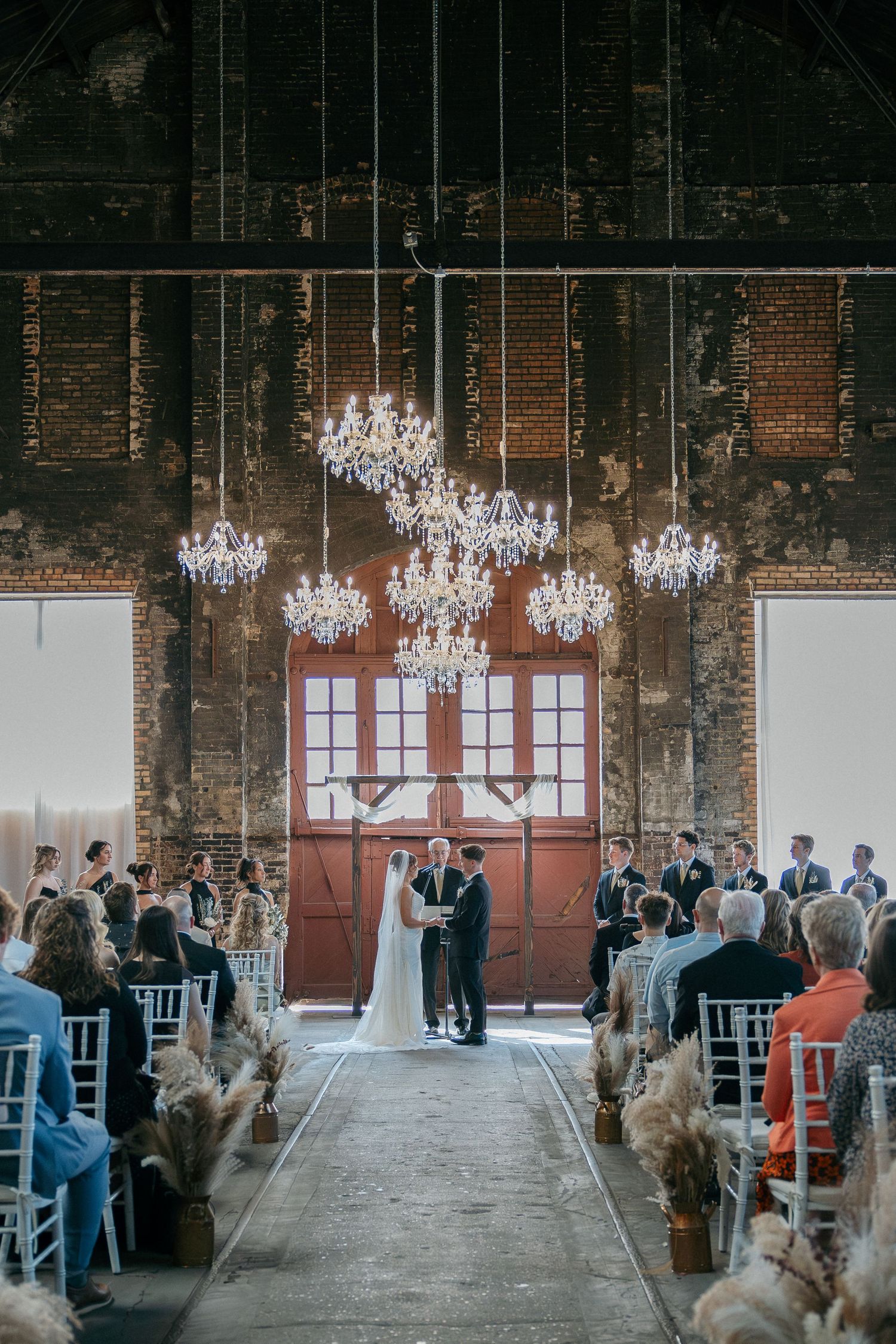 Bride and groom exchange vows under crystal chandeliers in a rustic brick wedding venue with white chairs.