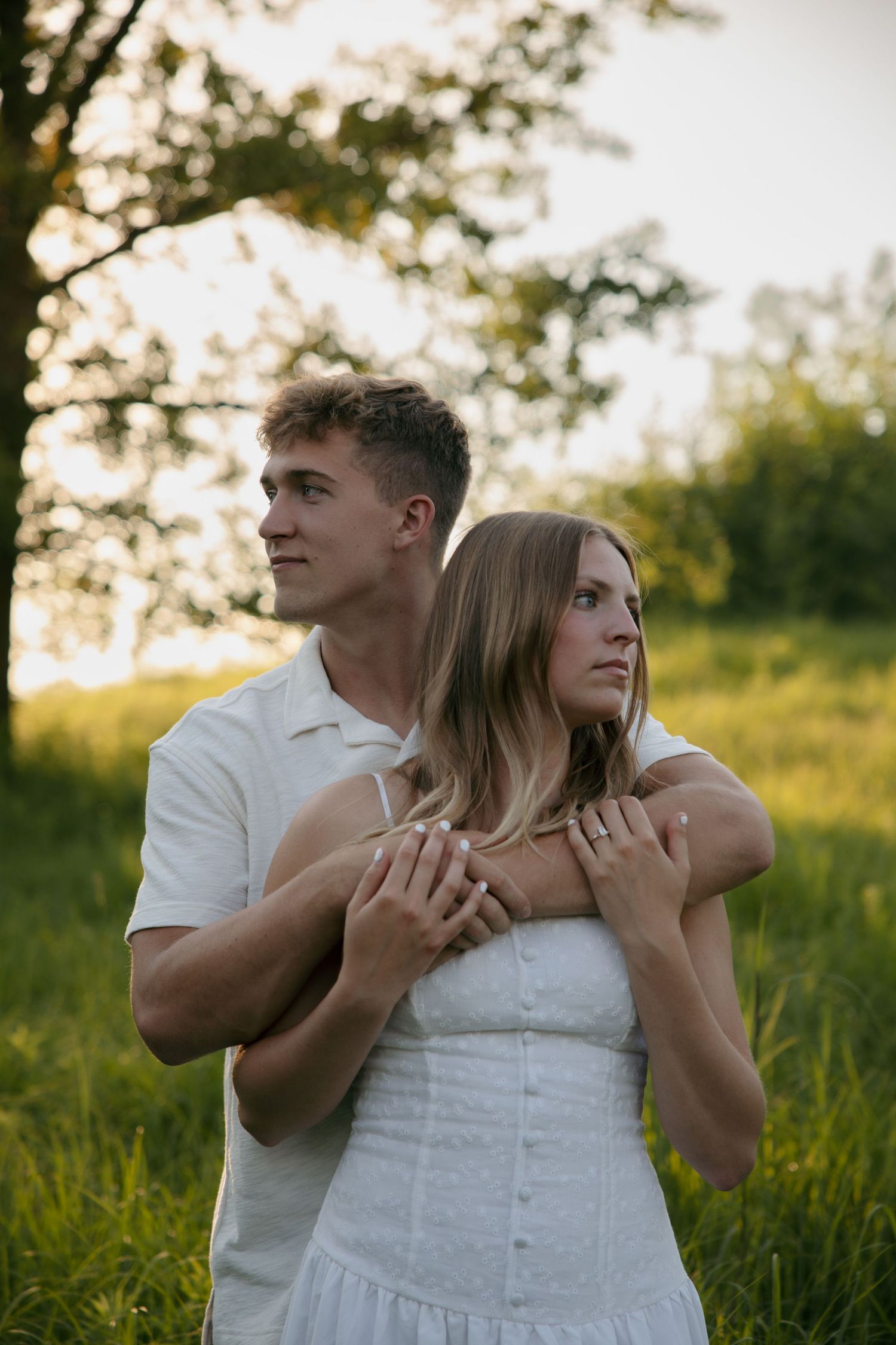 Man in white shirt embracing woman in white dress from behind in a golden sunset field.