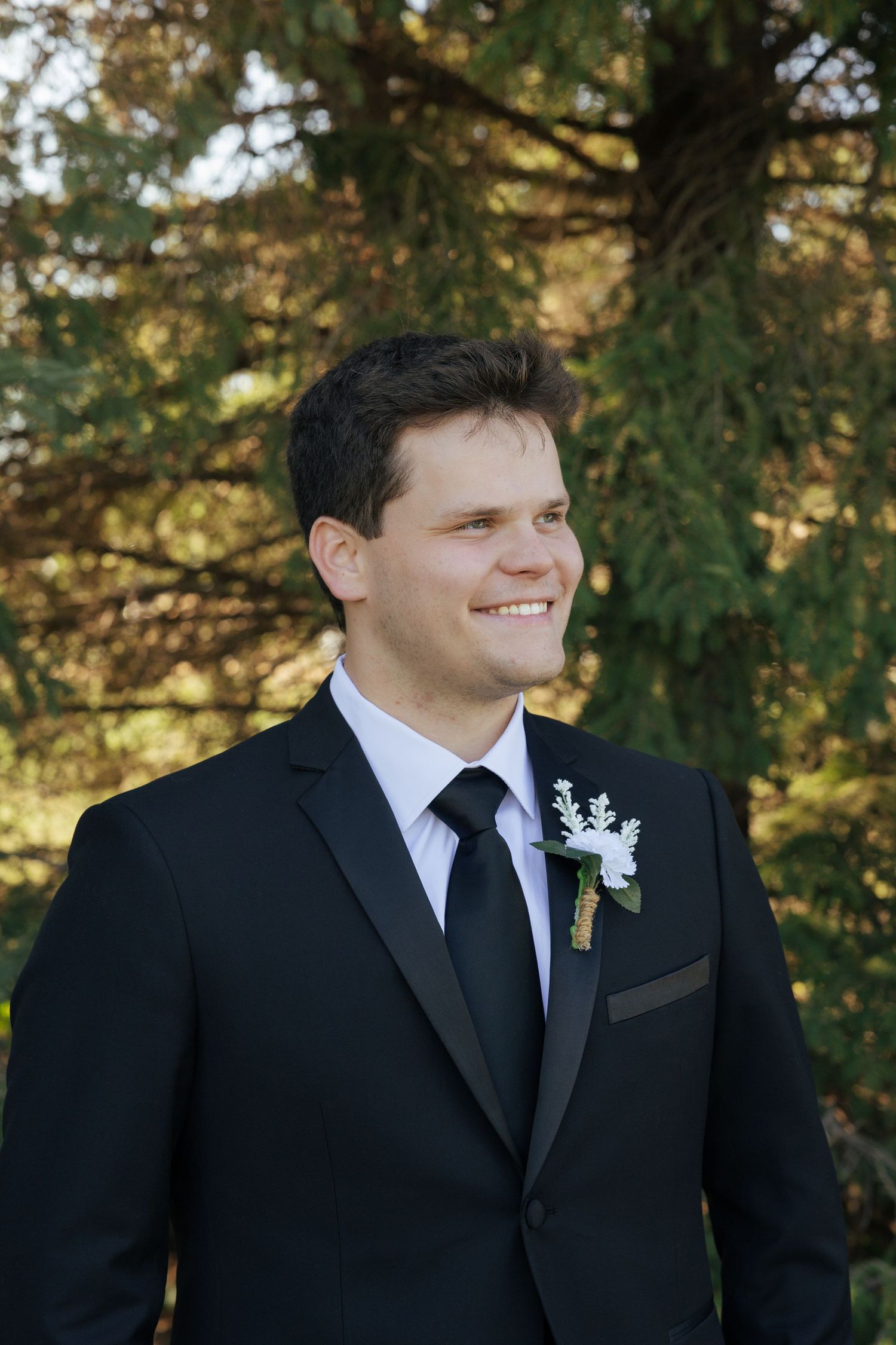 Smiling groom in black suit with boutonniere standing outdoors among evergreen trees.