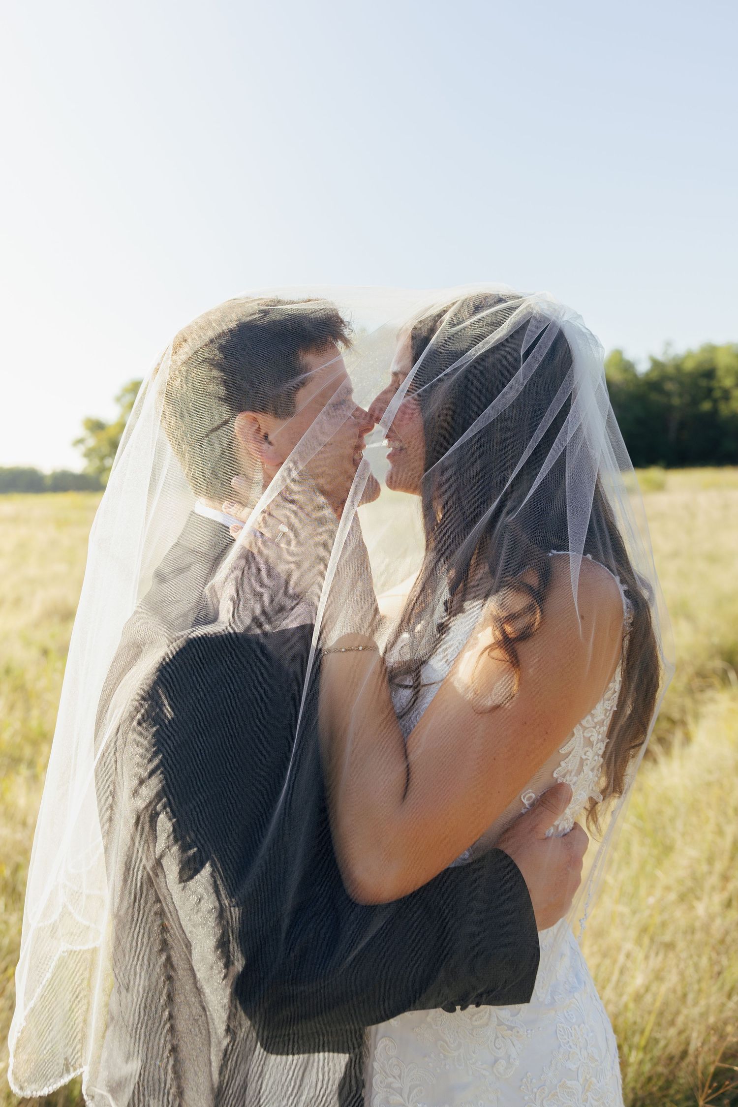 Bride and groom embracing under wedding veil in golden sunlit field on wedding day.