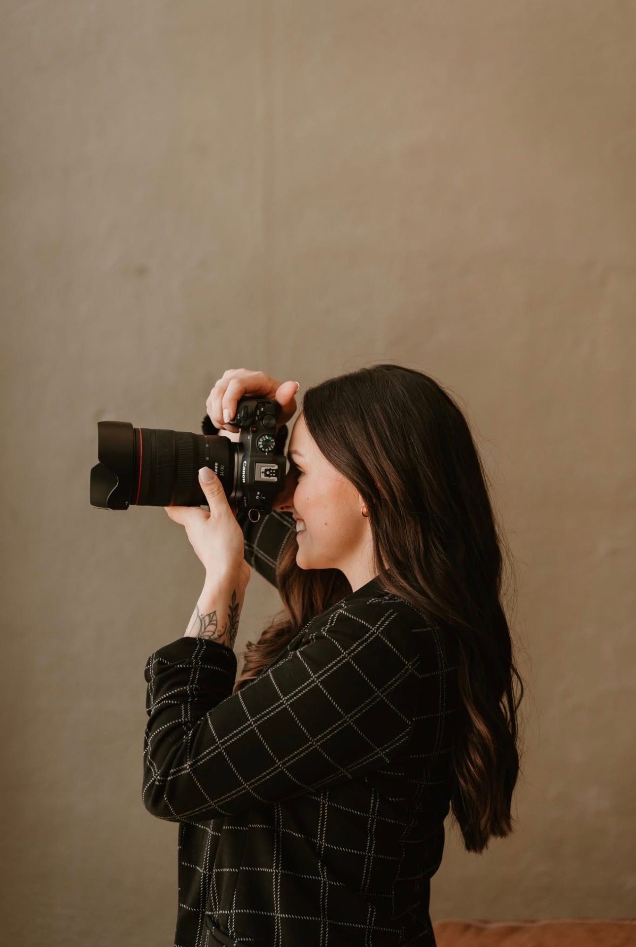 A woman with long dark hair photographs with a DSLR camera, wearing a plaid shirt, with a tattoo on her arm.