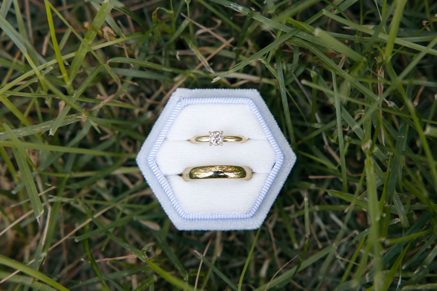 Two gold wedding rings and a diamond engagement ring displayed in a white hexagonal ring box on green grass.