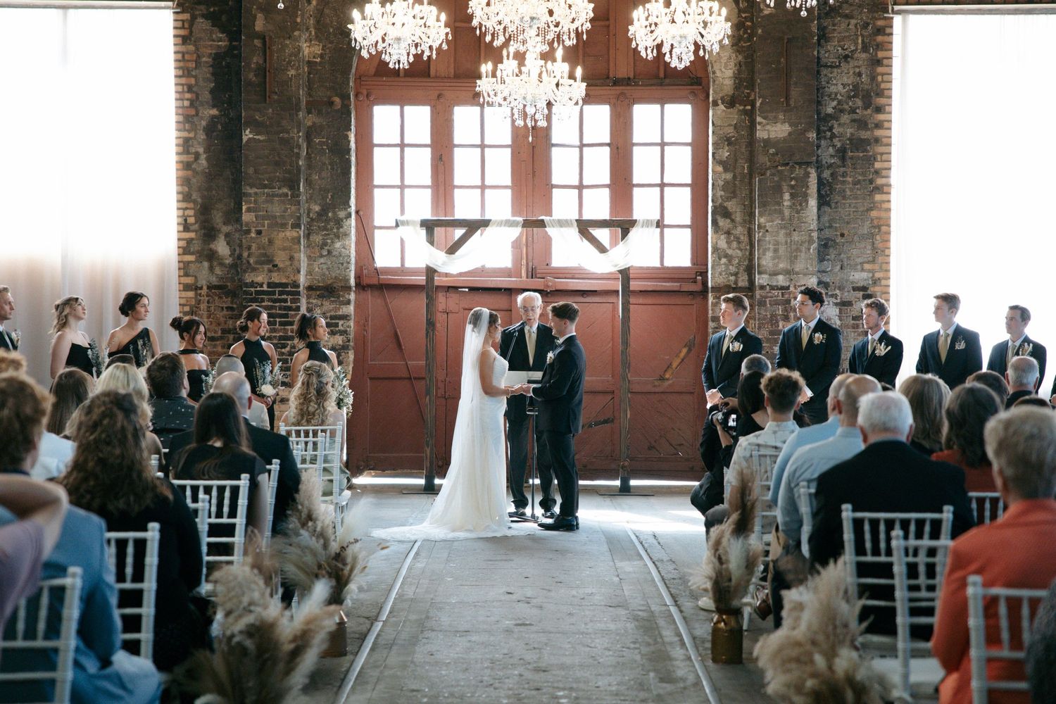 Elegant wedding ceremony in a rustic brick venue with chandeliers, red barn doors, and guests seated in white chairs.