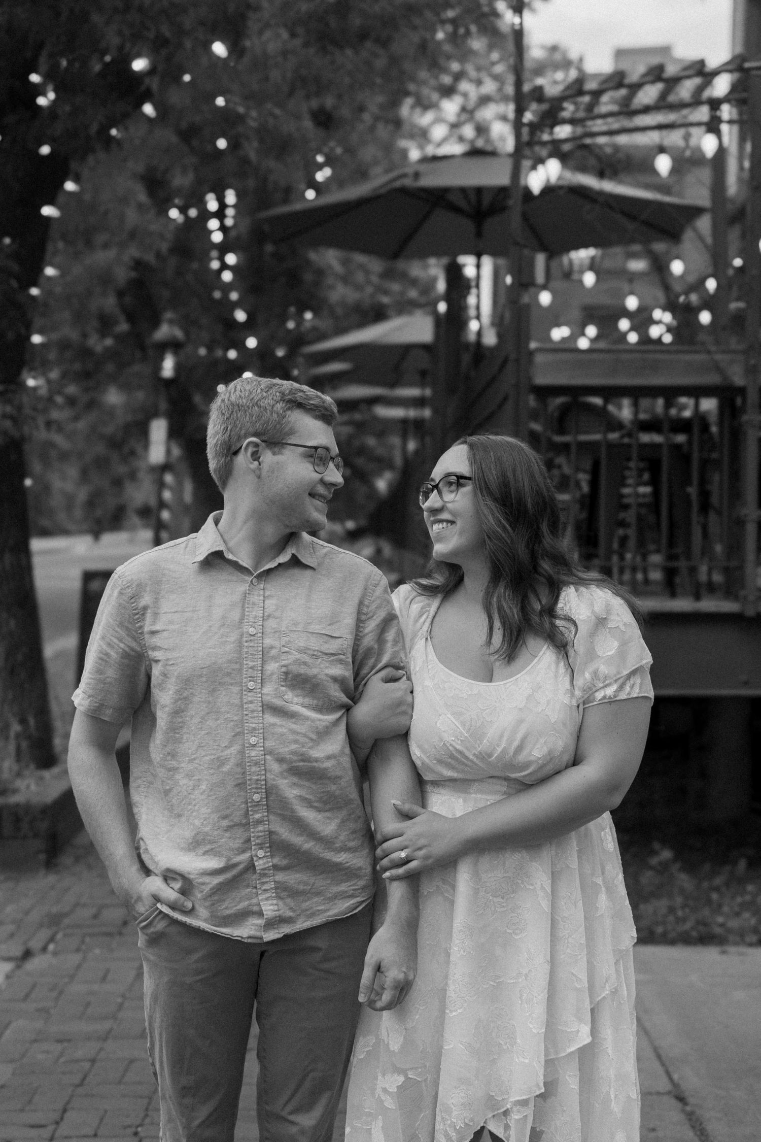Black and white photo of a smiling couple walking together outdoors on a brick path with string lights in the background.