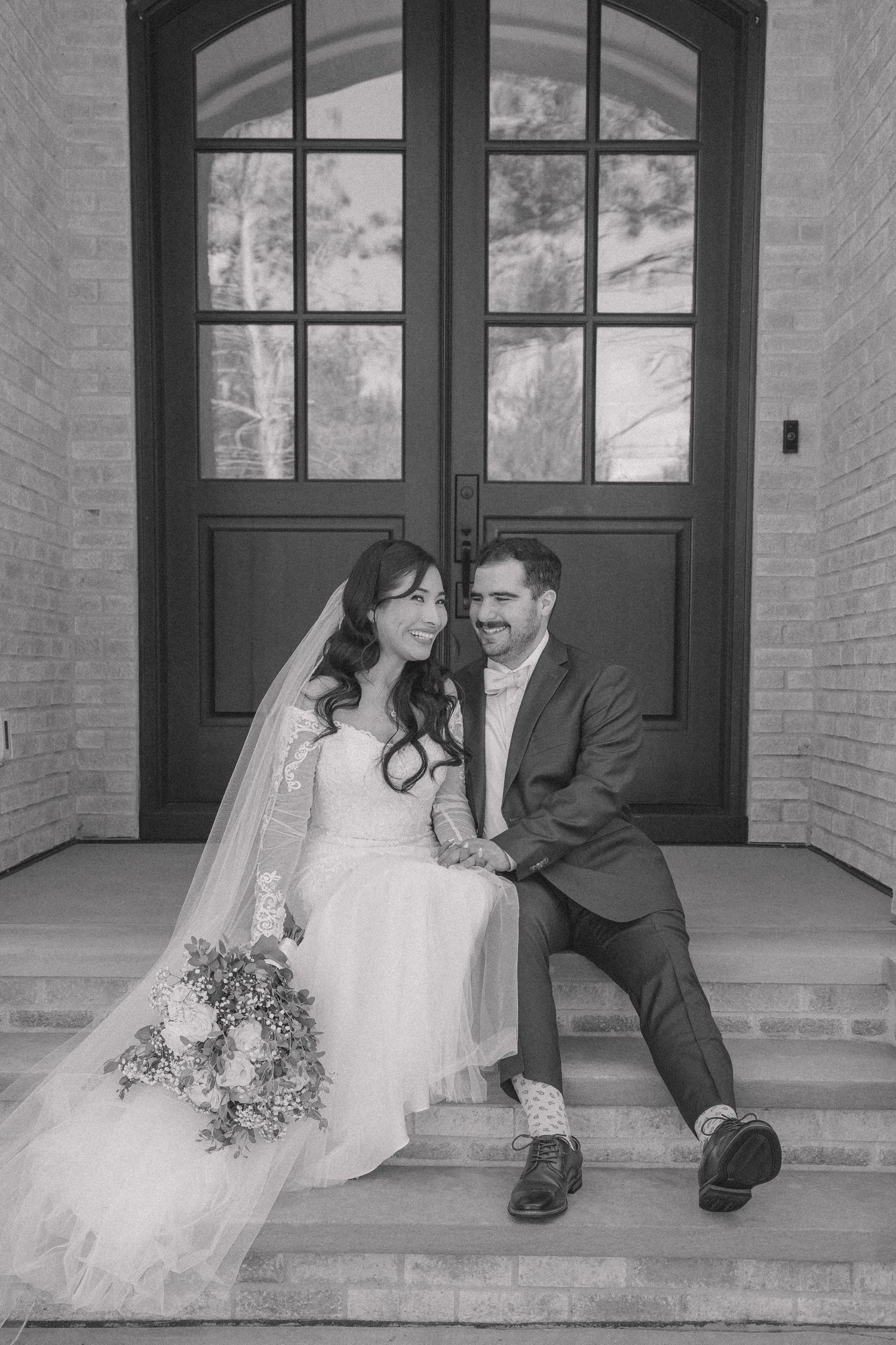 Black and white photo of a smiling bride and groom sitting on steps in front of elegant double doors on their wedding day.