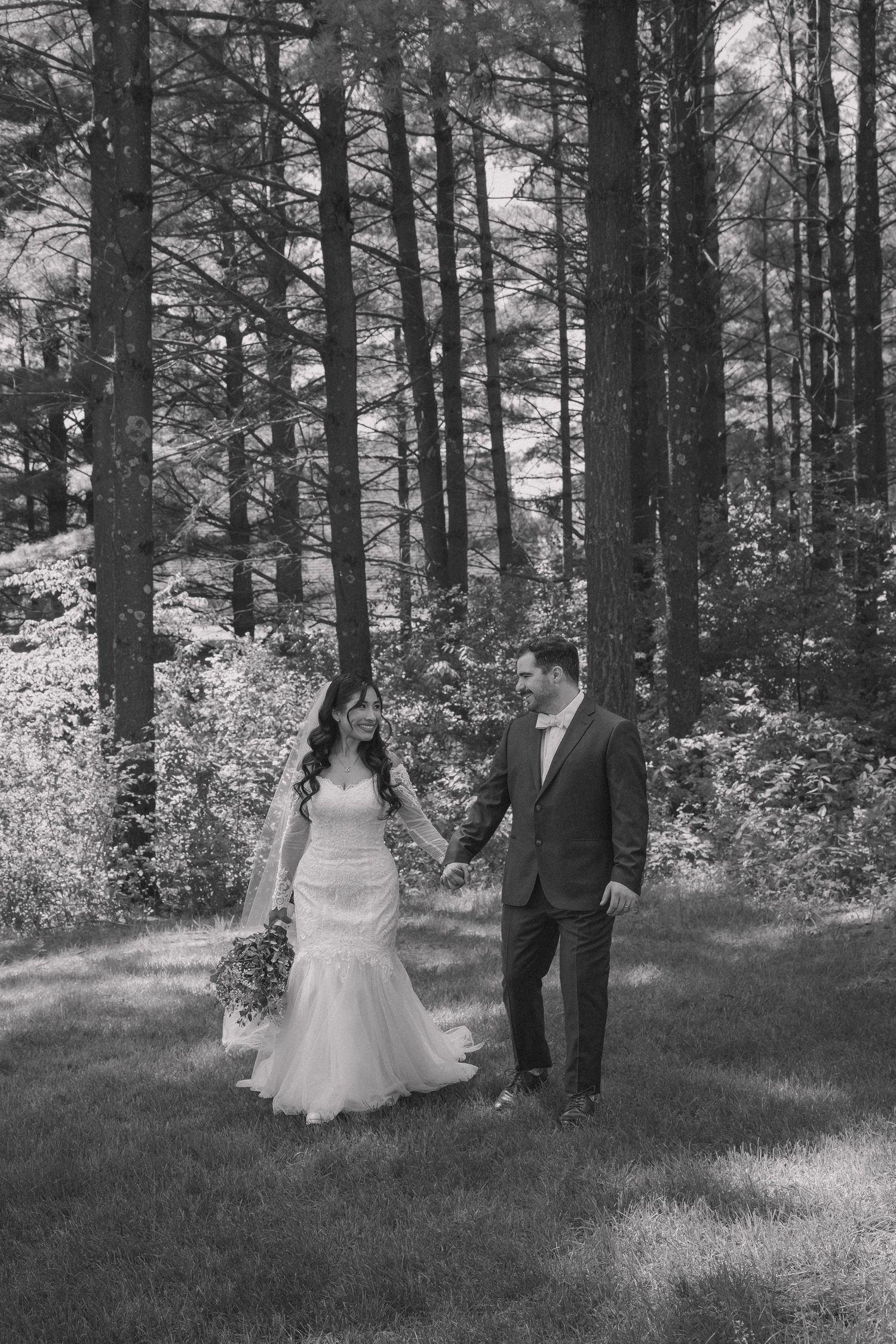 A bride and groom hold hands walking through a forest in a black and white wedding photo.