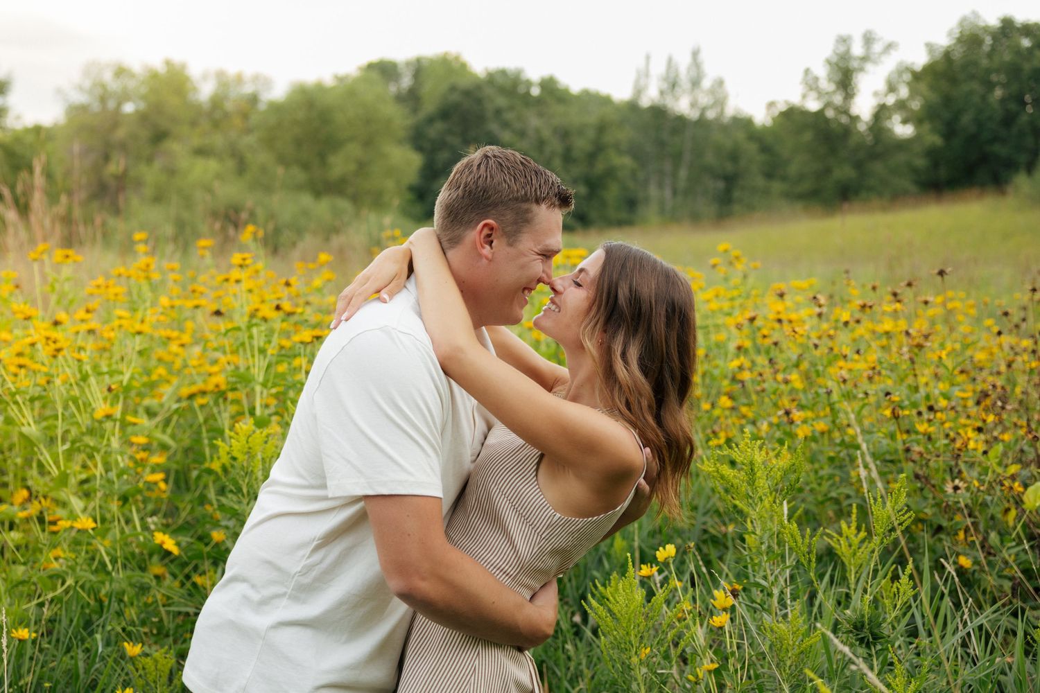 Couple embracing and almost kissing in a vibrant yellow wildflower field with lush green trees in the background.