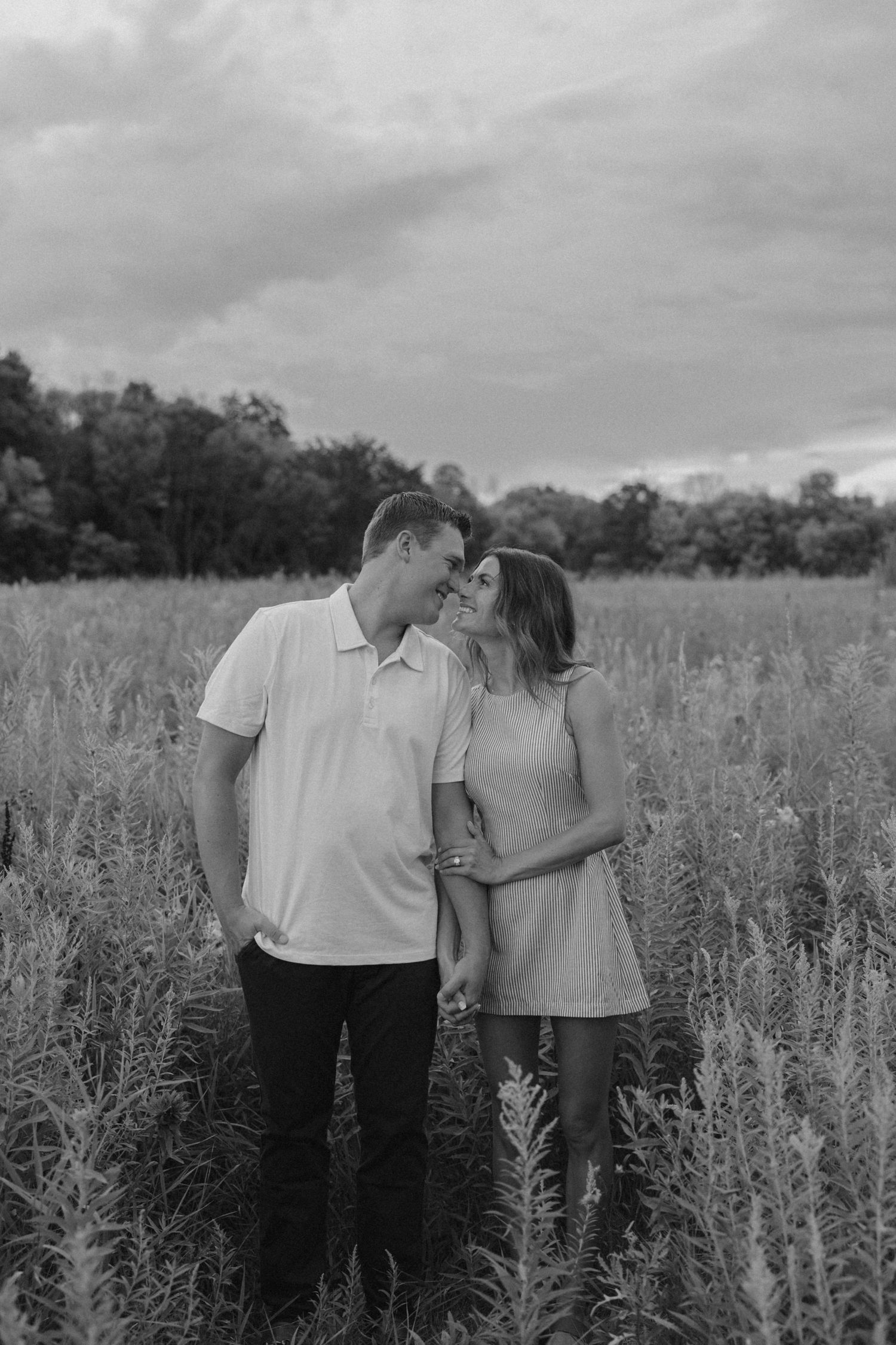 Black and white photo of a couple holding hands and smiling at each other in a lush field with trees behind.