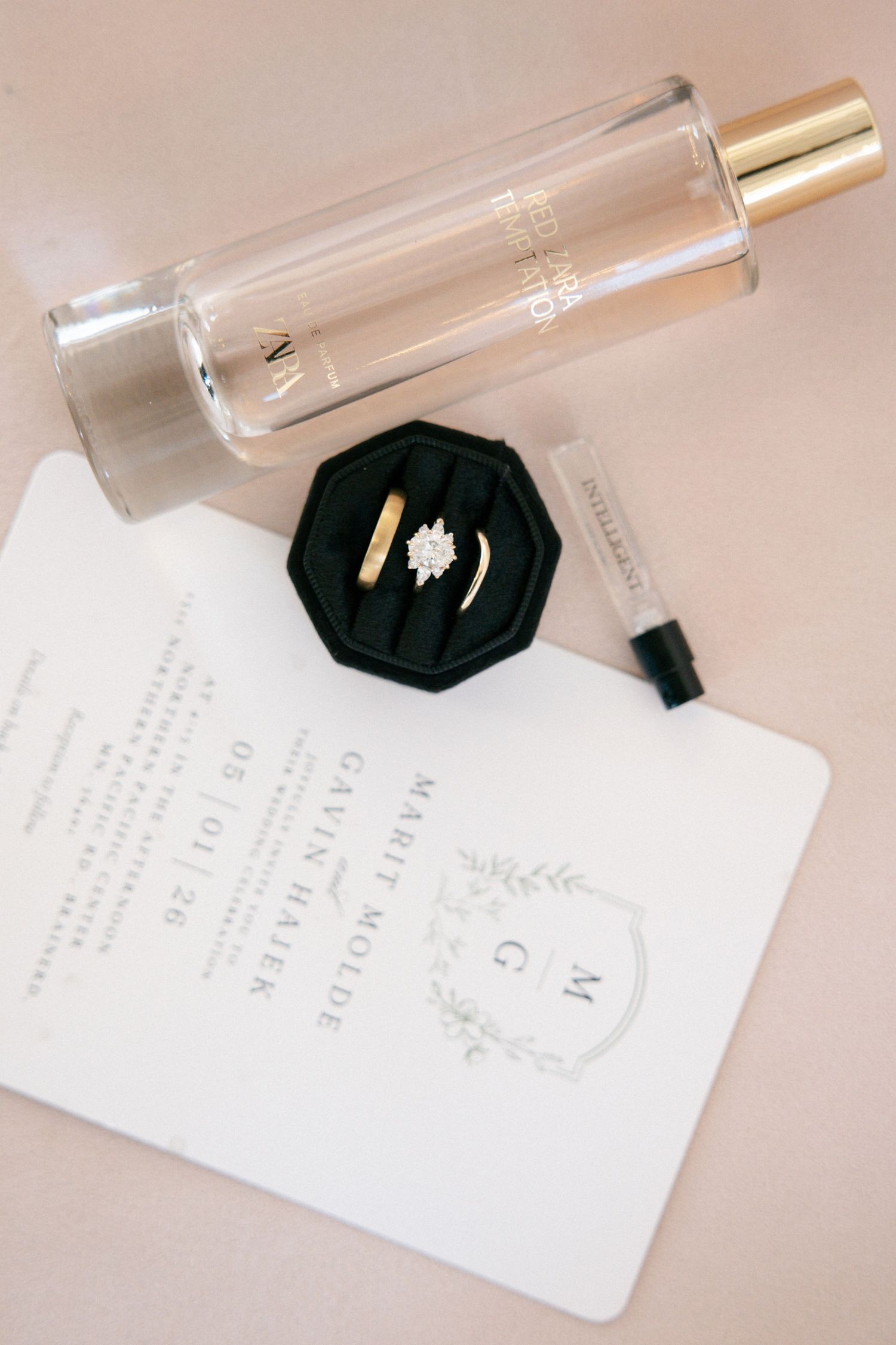 Wedding flat lay with diamond engagement ring in green velvet box, gold band, invitation, and perfume bottle.