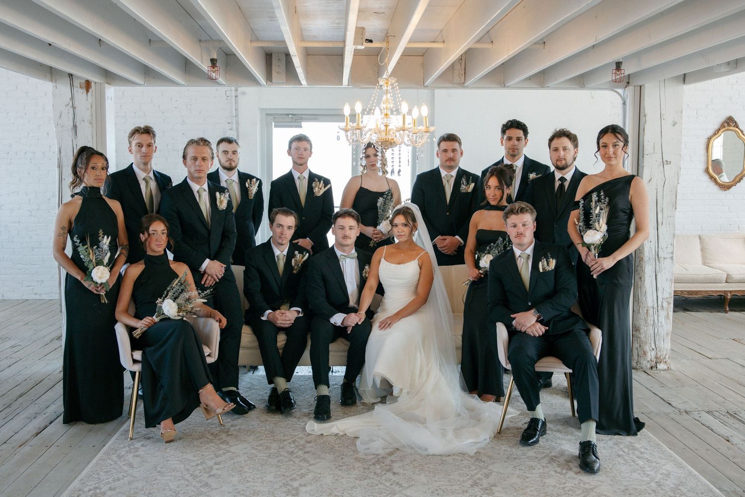 Large wedding party in black attire poses together in an elegant industrial venue with chandelier lighting.