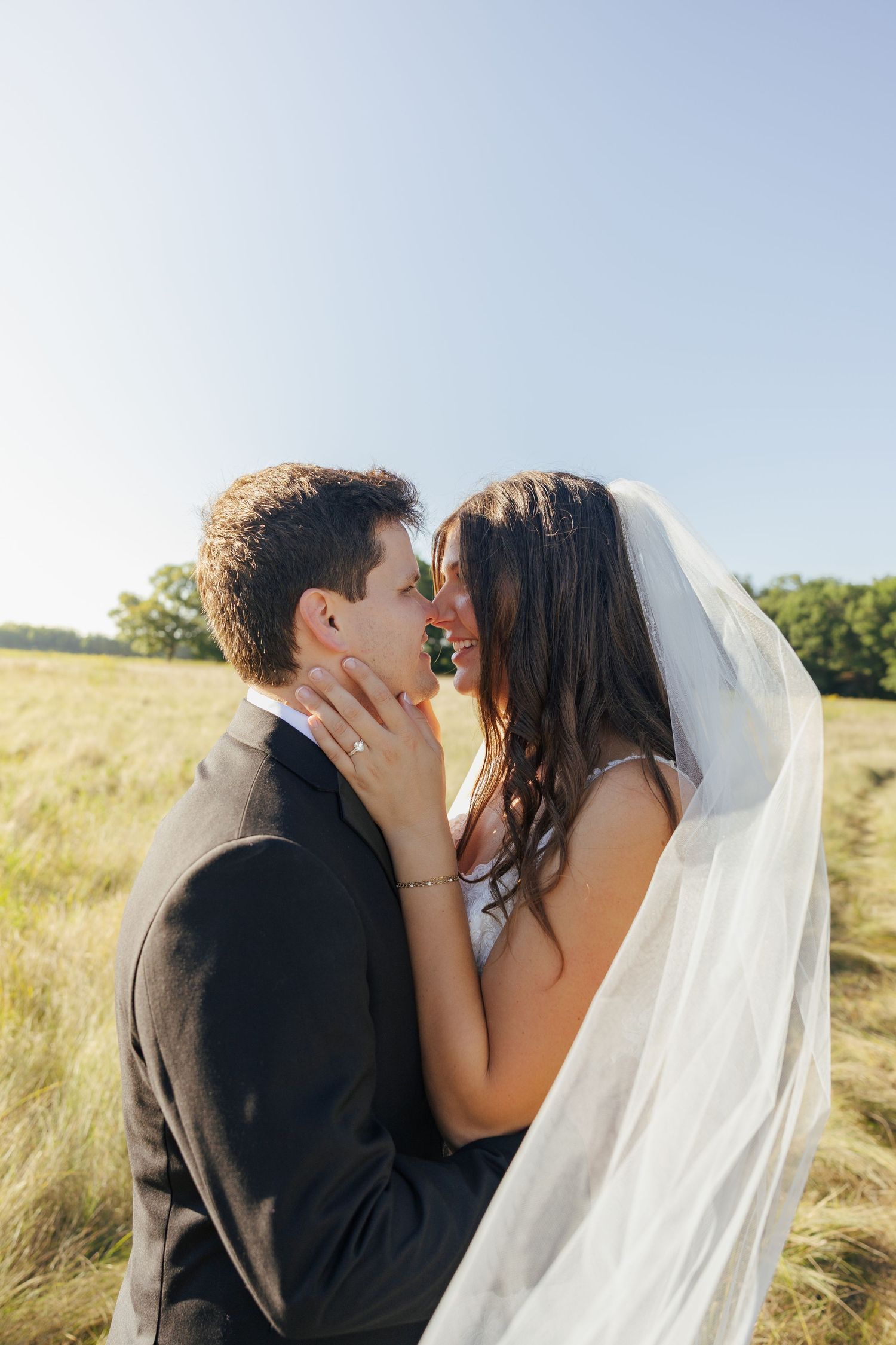 Bride and groom sharing romantic moment under flowing veil in sunny grassy field.