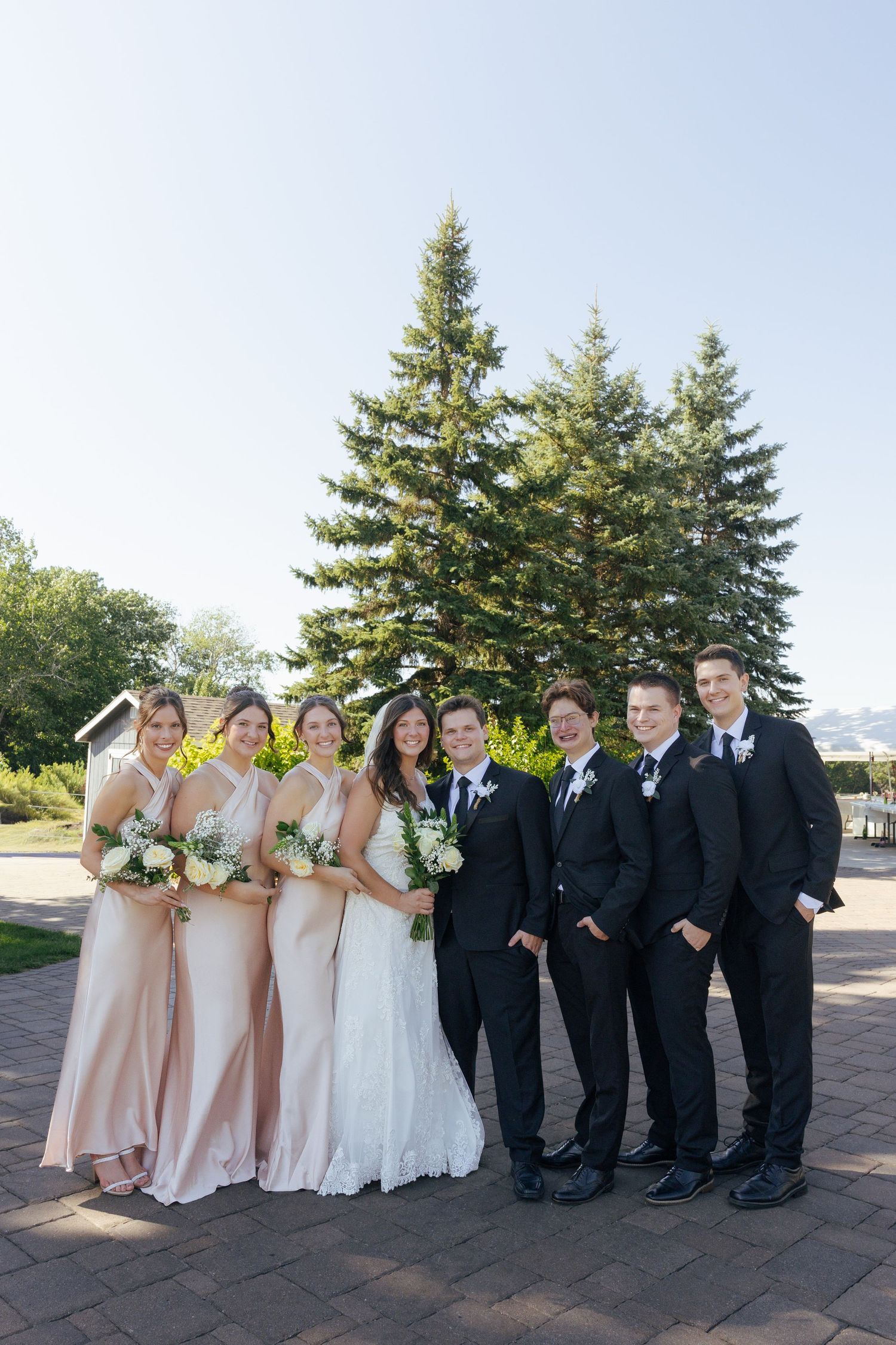 Wedding party posing outdoors with bridesmaids in blush gowns and groomsmen in black suits before tall evergreen trees.