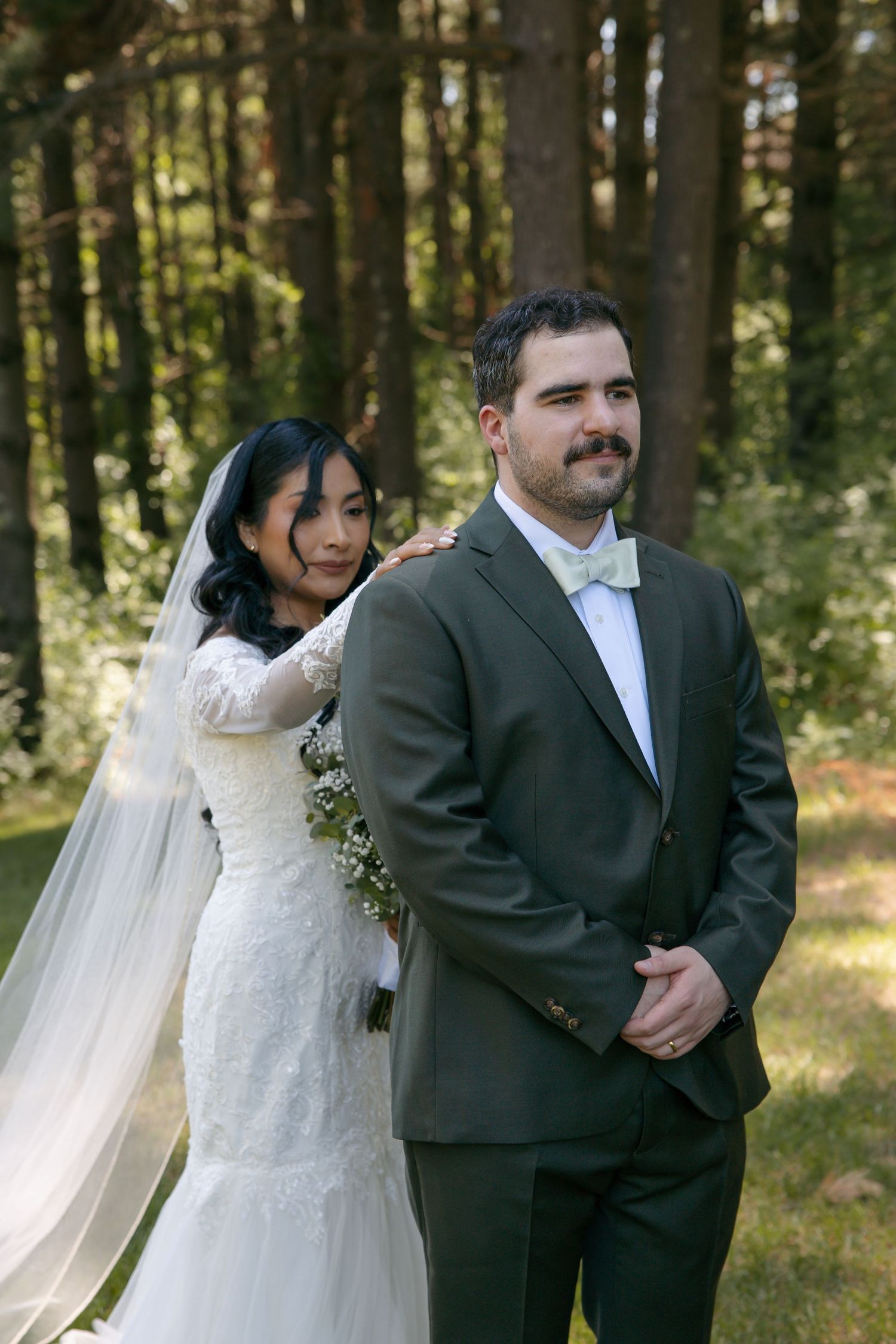 Bride in white lace gown and veil stands behind groom in olive suit with bow tie during outdoor forest wedding.