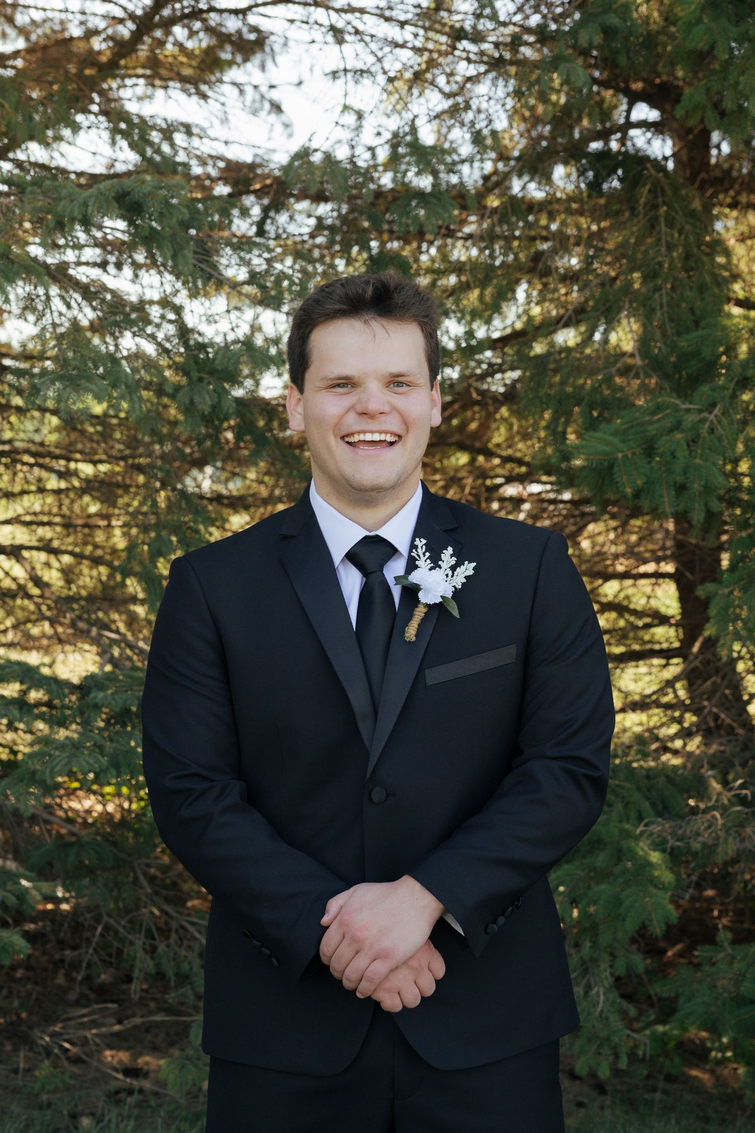 Smiling groom in black tuxedo with white boutonniere stands outdoors among green pine trees on wedding day.