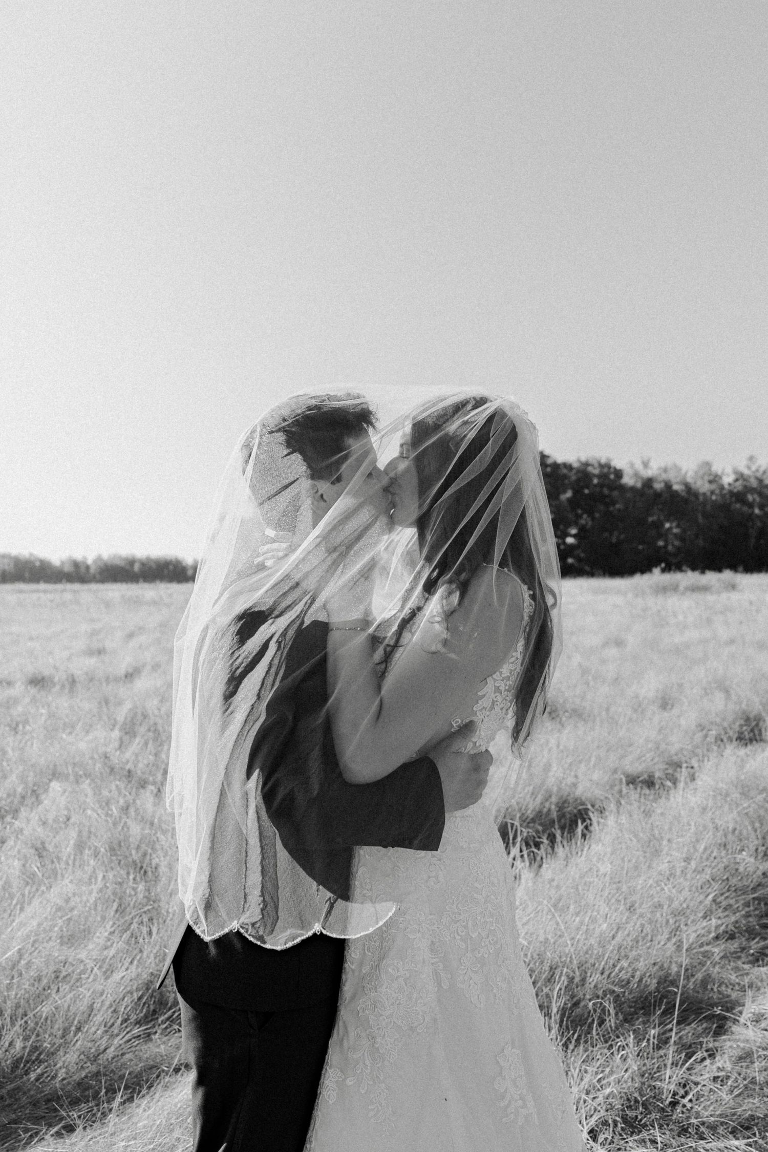 Black and white wedding photo of couple kissing beneath flowing bridal veil in field.