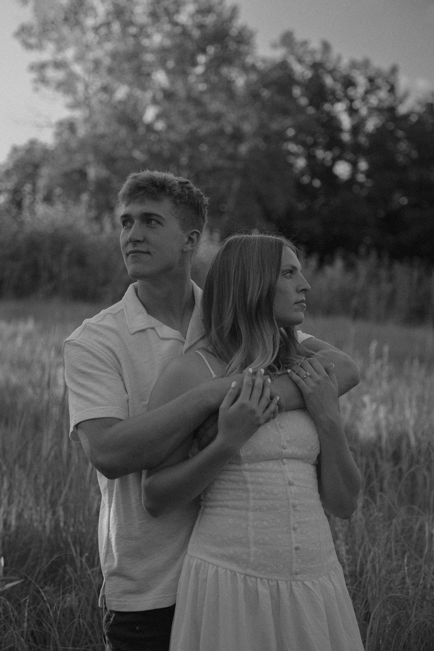 Black and white photo of a young couple embracing in a field, man holding woman from behind among tall grass.
