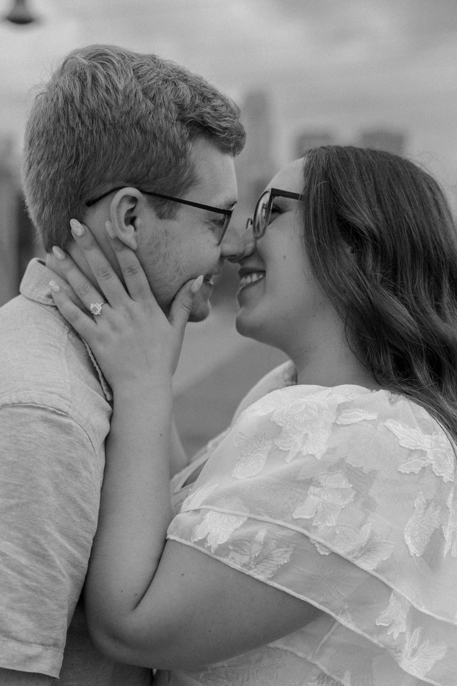 Black and white photo of a smiling couple in glasses sharing a romantic moment, woman wearing engagement ring.