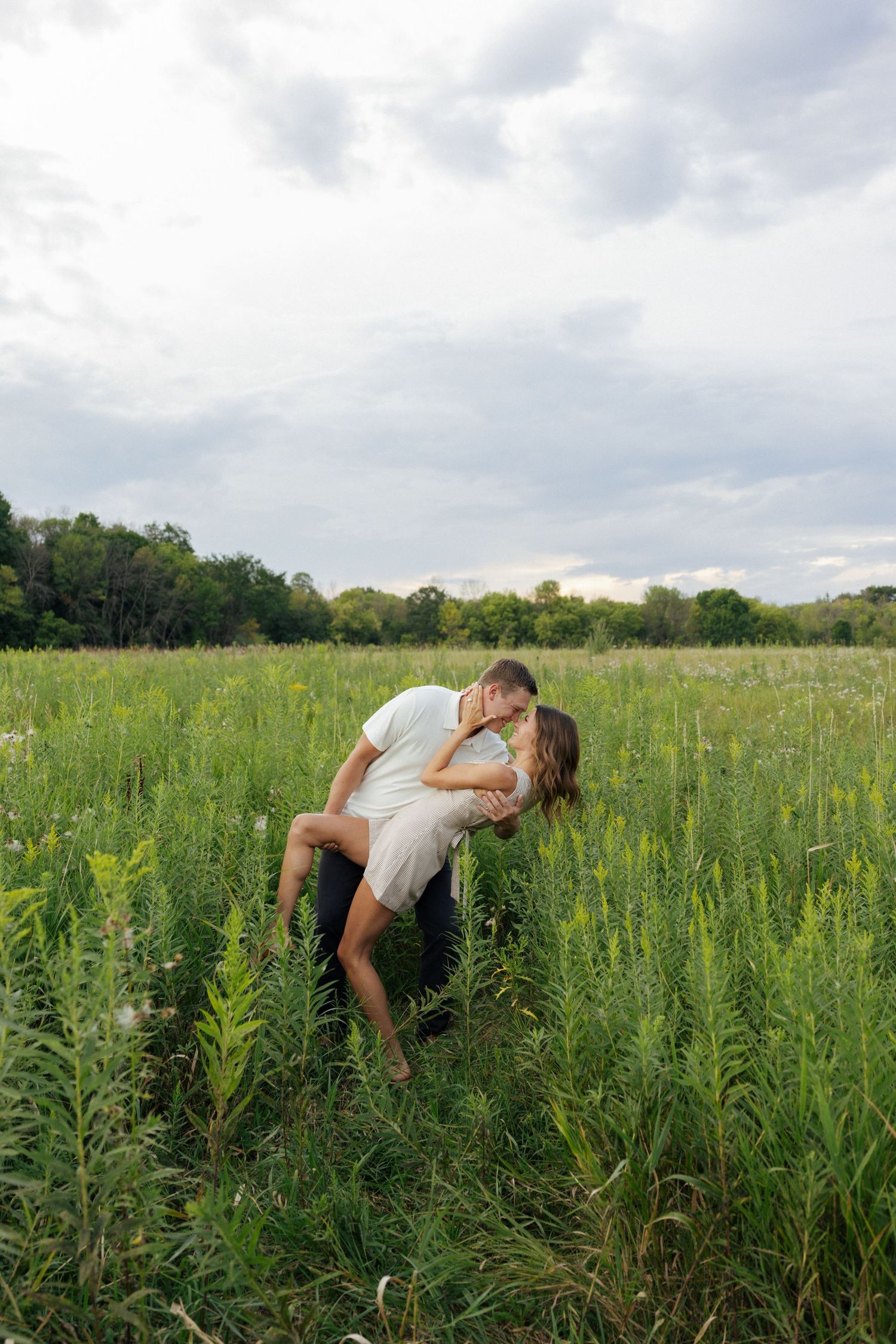 Couple sharing a romantic dip kiss surrounded by tall green wildflowers in an open field.