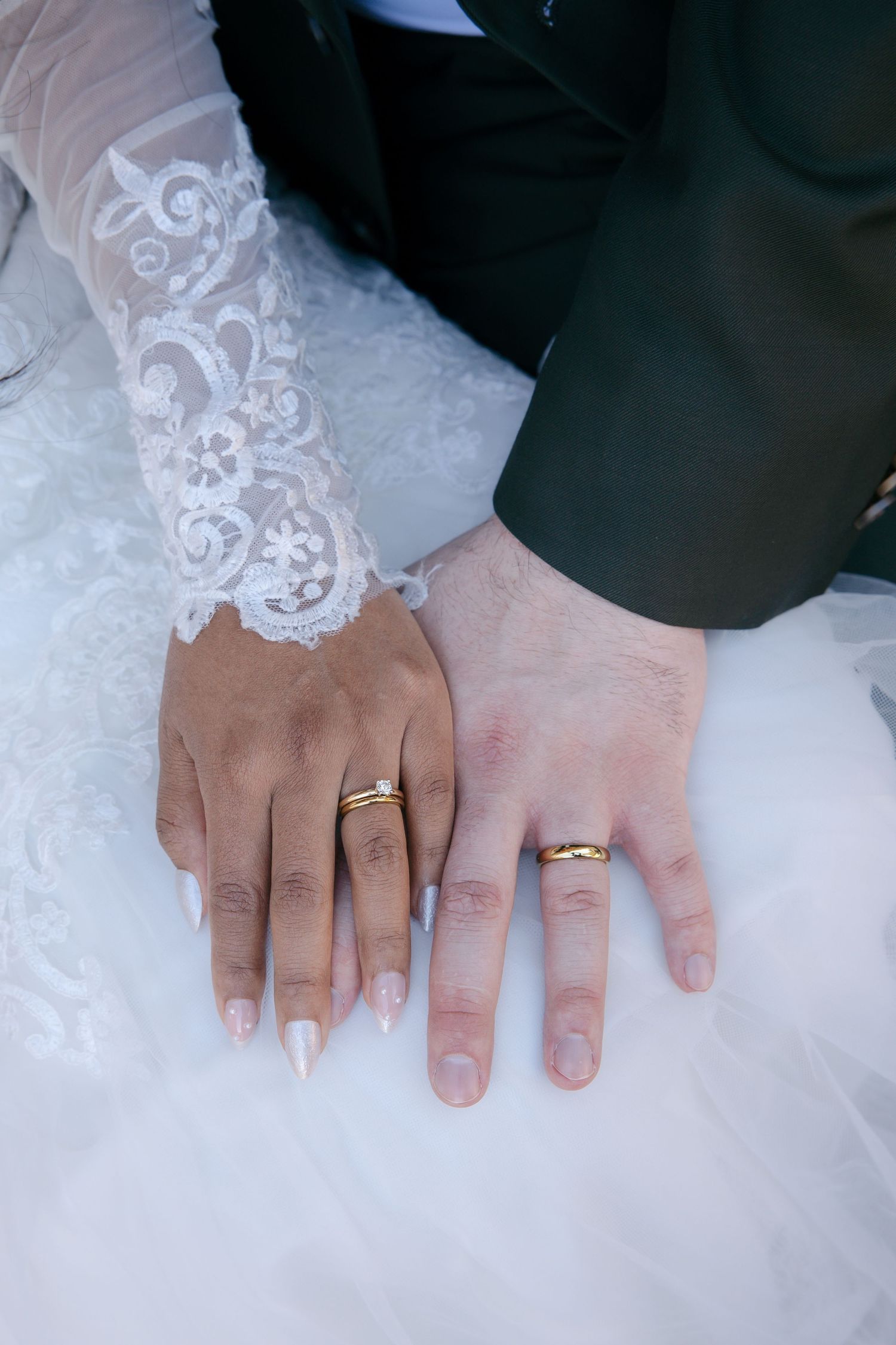 Bride and groom displaying gold wedding rings on their hands, with lace bridal gown sleeve visible.