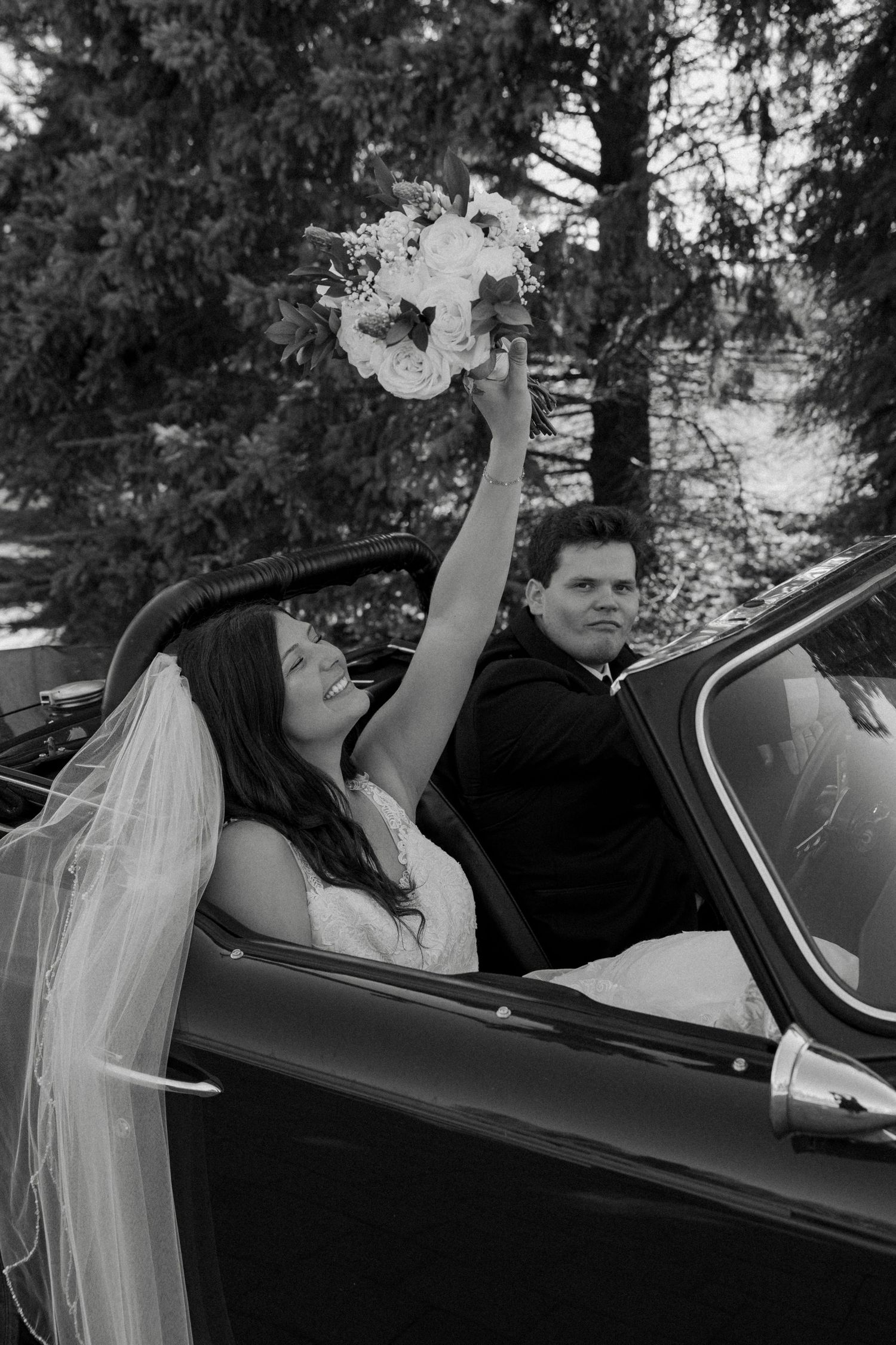 Black and white photo of a bride raising her bouquet joyfully while sitting in a convertible car with the groom.