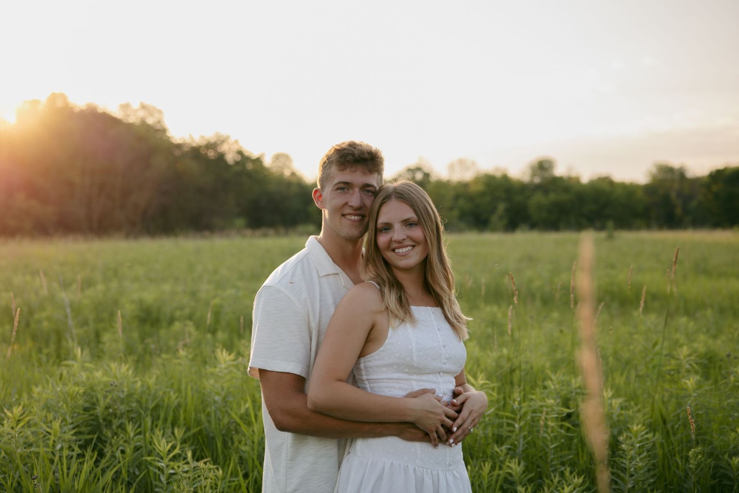 Smiling couple posing together in green field at sunset, both wearing white in a warm golden light.