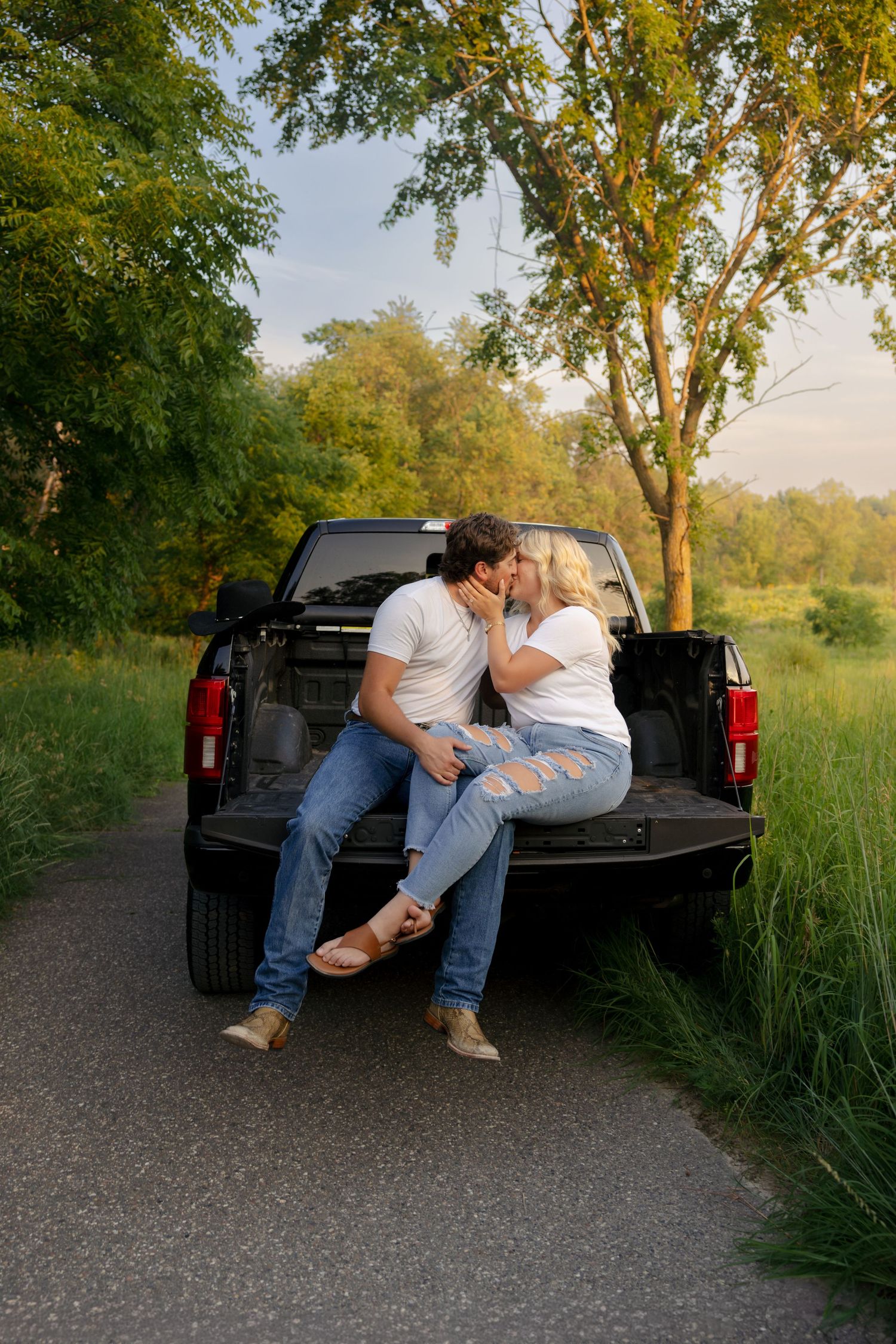 Couple kissing on the tailgate of a black pickup truck surrounded by lush green trees at golden hour.