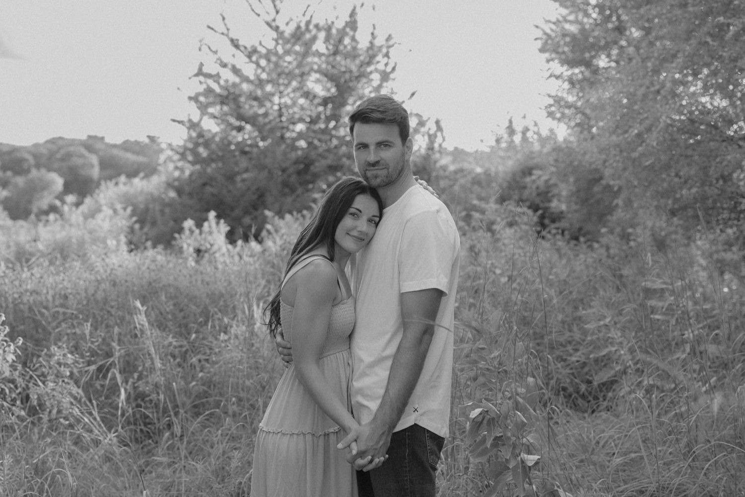 Black and white photo of a couple embracing in a lush outdoor field, holding hands and smiling softly.