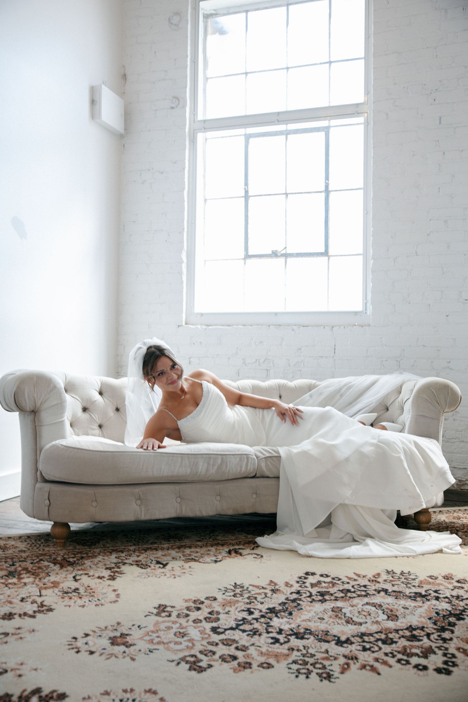 Bride in white wedding gown and veil reclining gracefully on a tufted ivory sofa in a bright, airy room.