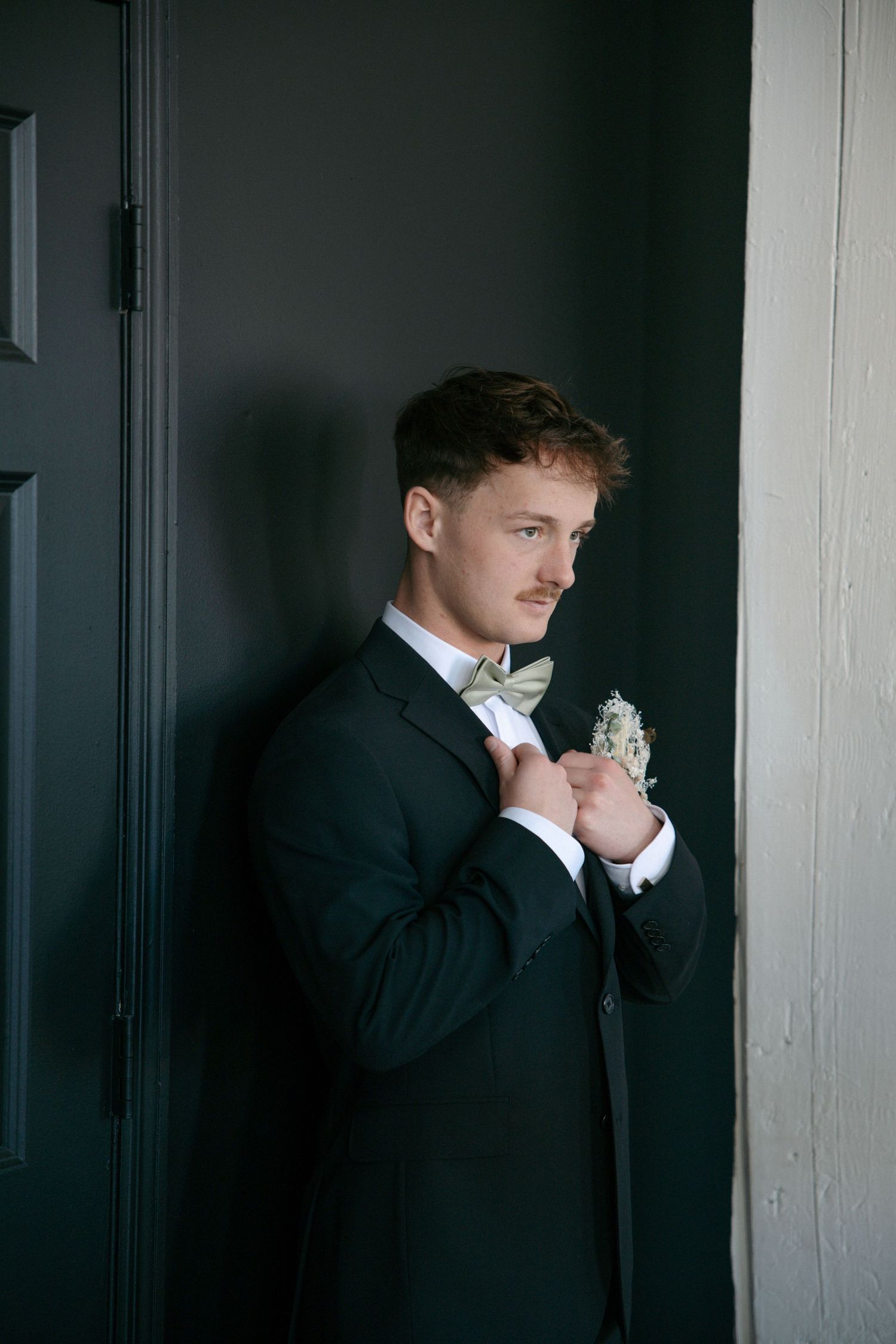 Dapper groom in black tuxedo adjusts his boutonniere while posing against a dark wall.