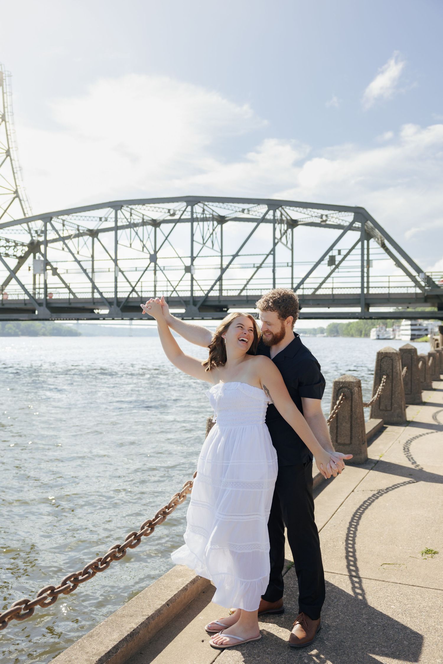 Couple dancing and laughing by a river with a steel bridge backdrop on a sunny day.