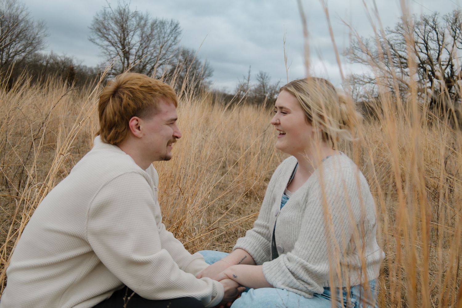A young couple sits together in a golden grass field, smiling and holding hands on a cloudy day.