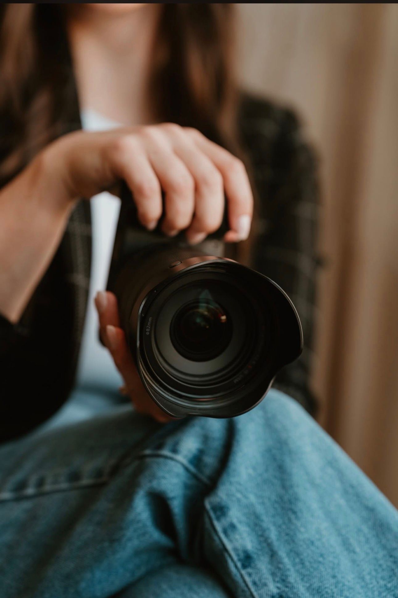 A photographer holds a professional DSLR camera with a wide-angle lens while sitting in jeans and a plaid shirt.