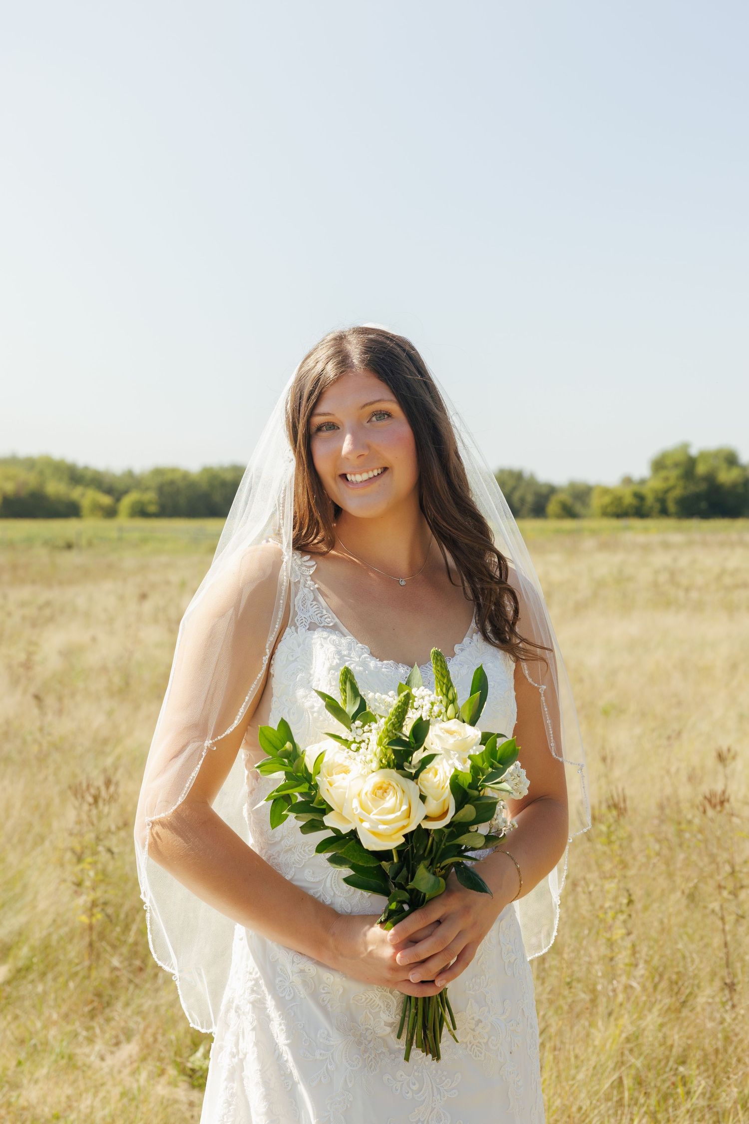 Bride in lace wedding gown and veil holding white rose bouquet smiling in sunlit outdoor field.