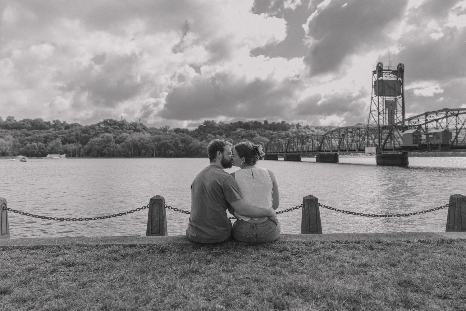 Couple sitting together on grass by a river with a historic bridge in the background, black and white photo.