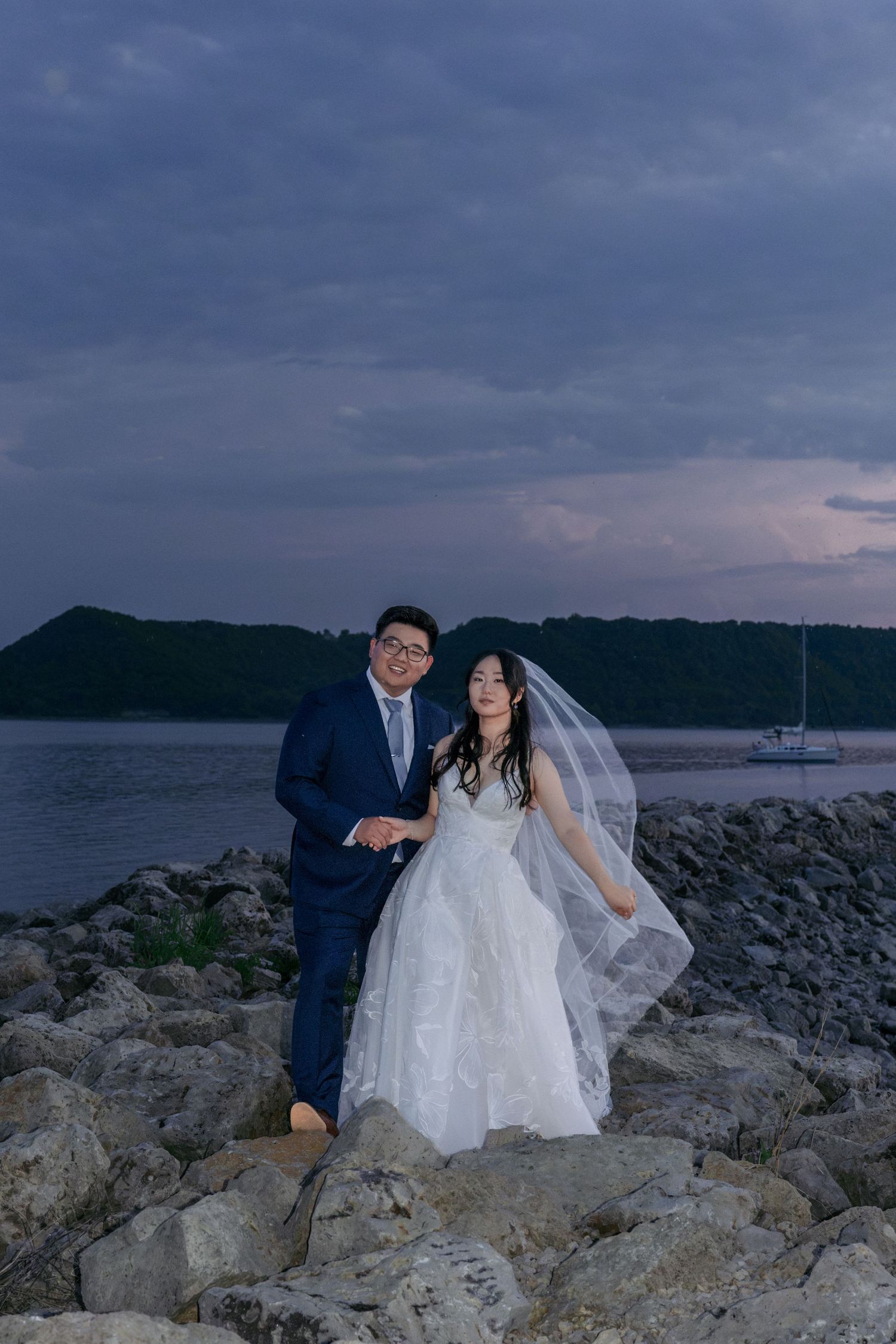 Bride and groom pose on rocky shoreline at twilight with dramatic cloudy skies and water in the background.