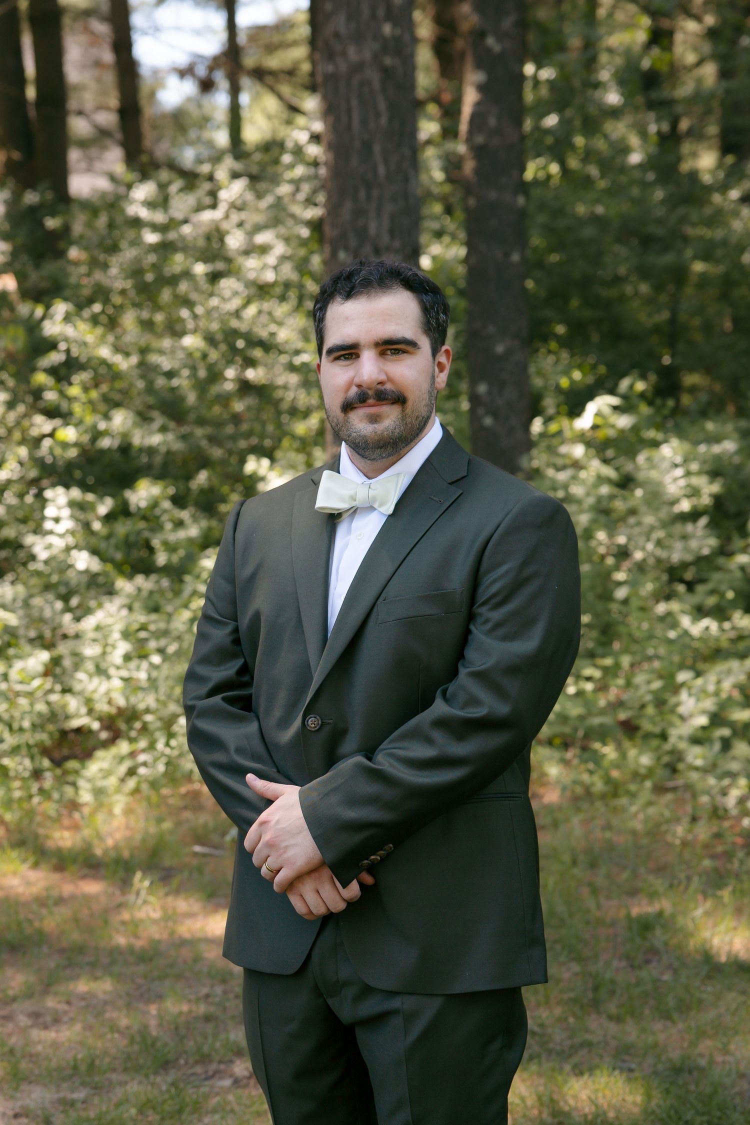 Groom in dark green suit with sage bow tie smiling outdoors in wooded setting.