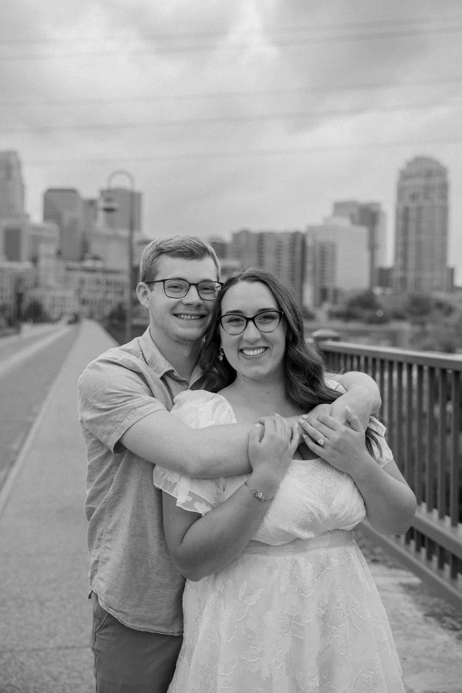 Black and white photo of a smiling couple embracing on a bridge with a city skyline backdrop.