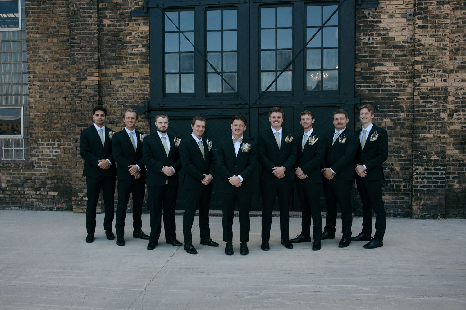 Groom and groomsmen in matching black suits with white boutonnieres posing outside a brick wedding venue.