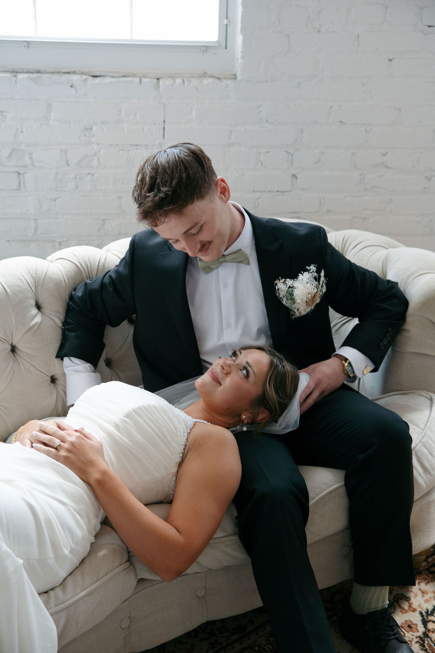 Bride in white gown reclines on tufted sofa while groom in dark suit smiles down at her in bright white brick room.