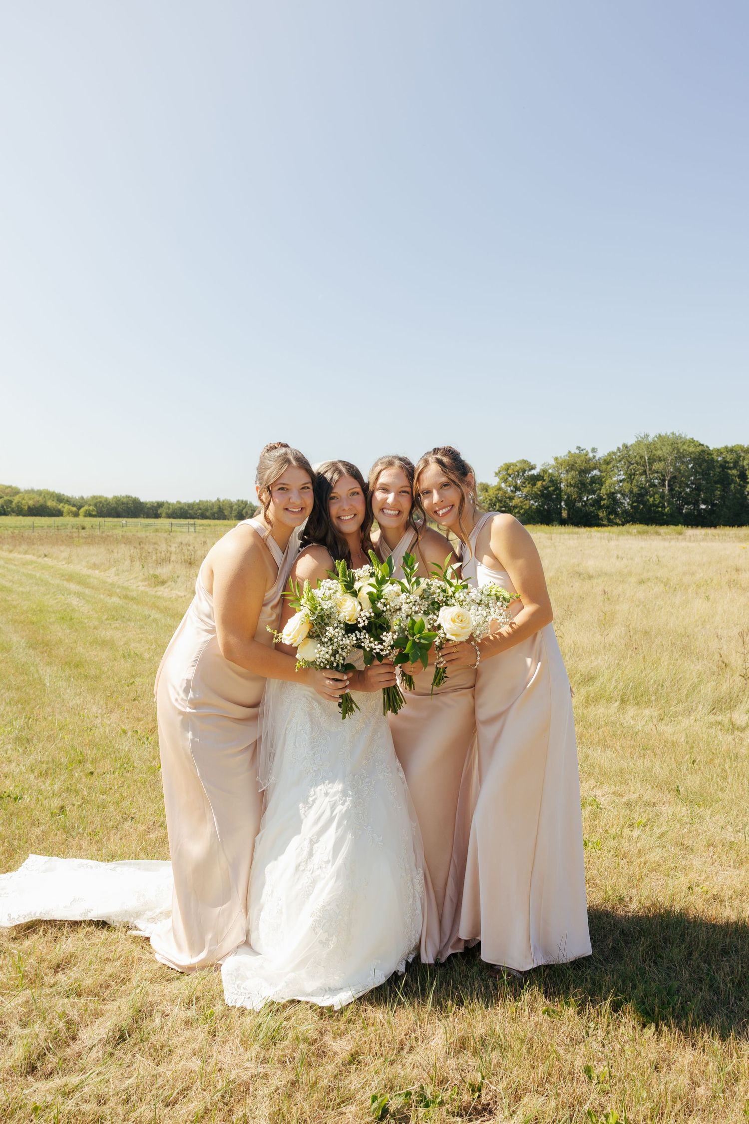 Bride and three bridesmaids in blush gowns holding white bouquets, smiling together in a sunny green field.