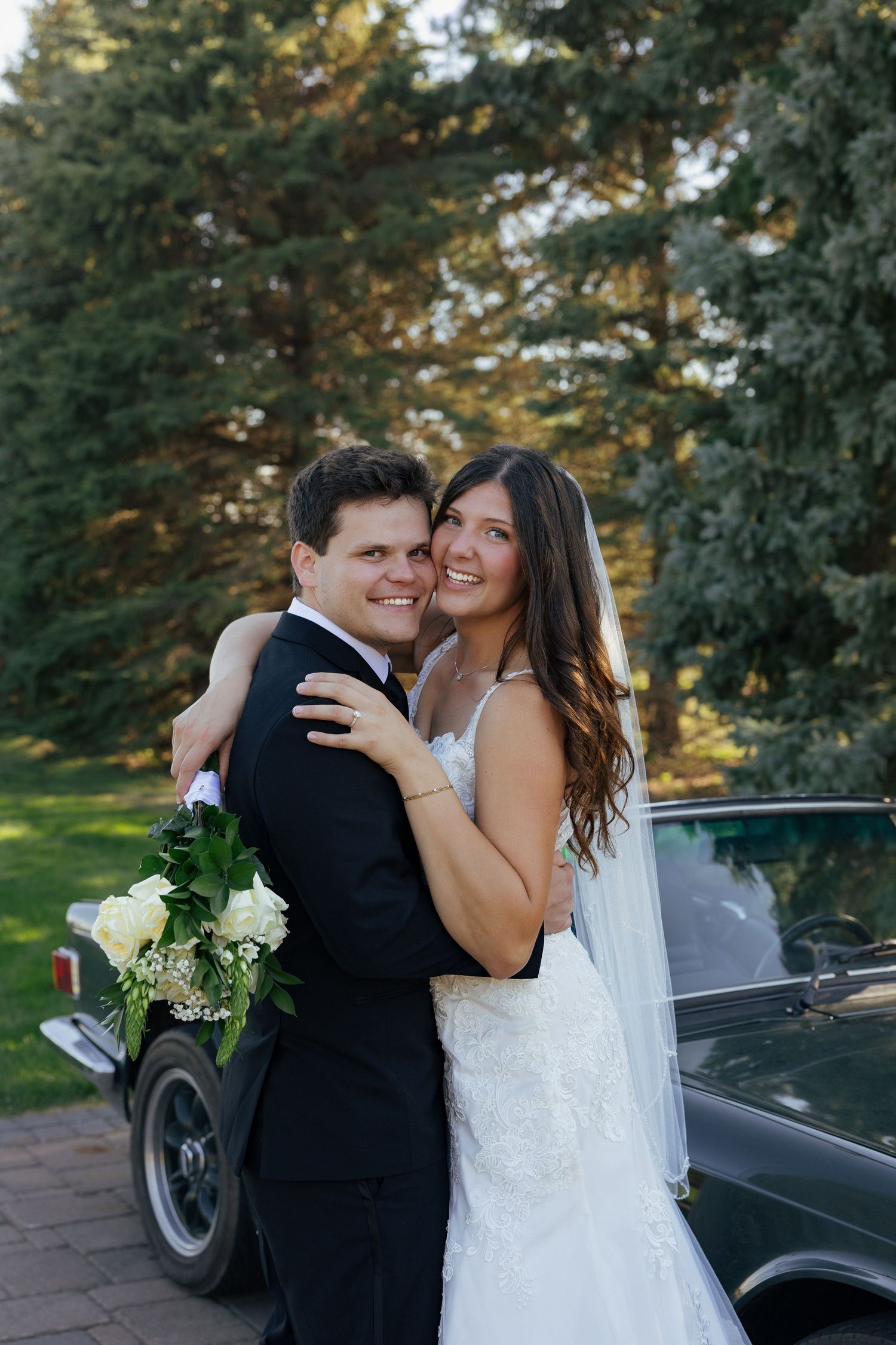 A smiling bride and groom embrace near a vintage car surrounded by tall evergreen trees on their wedding day.