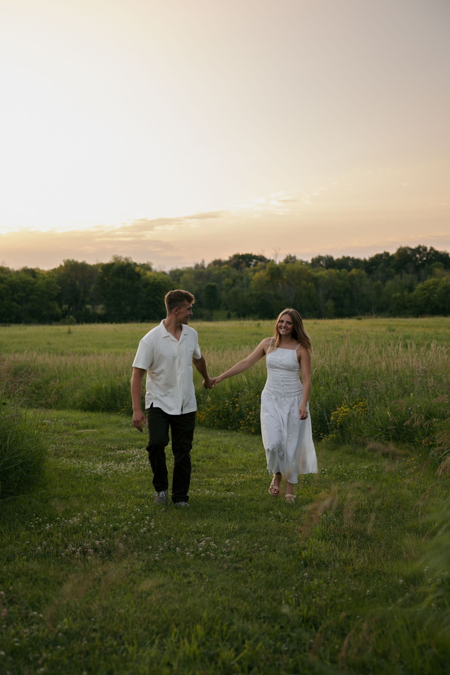 A couple holds hands walking through a lush green field at sunset, woman in white dress, man in white shirt.