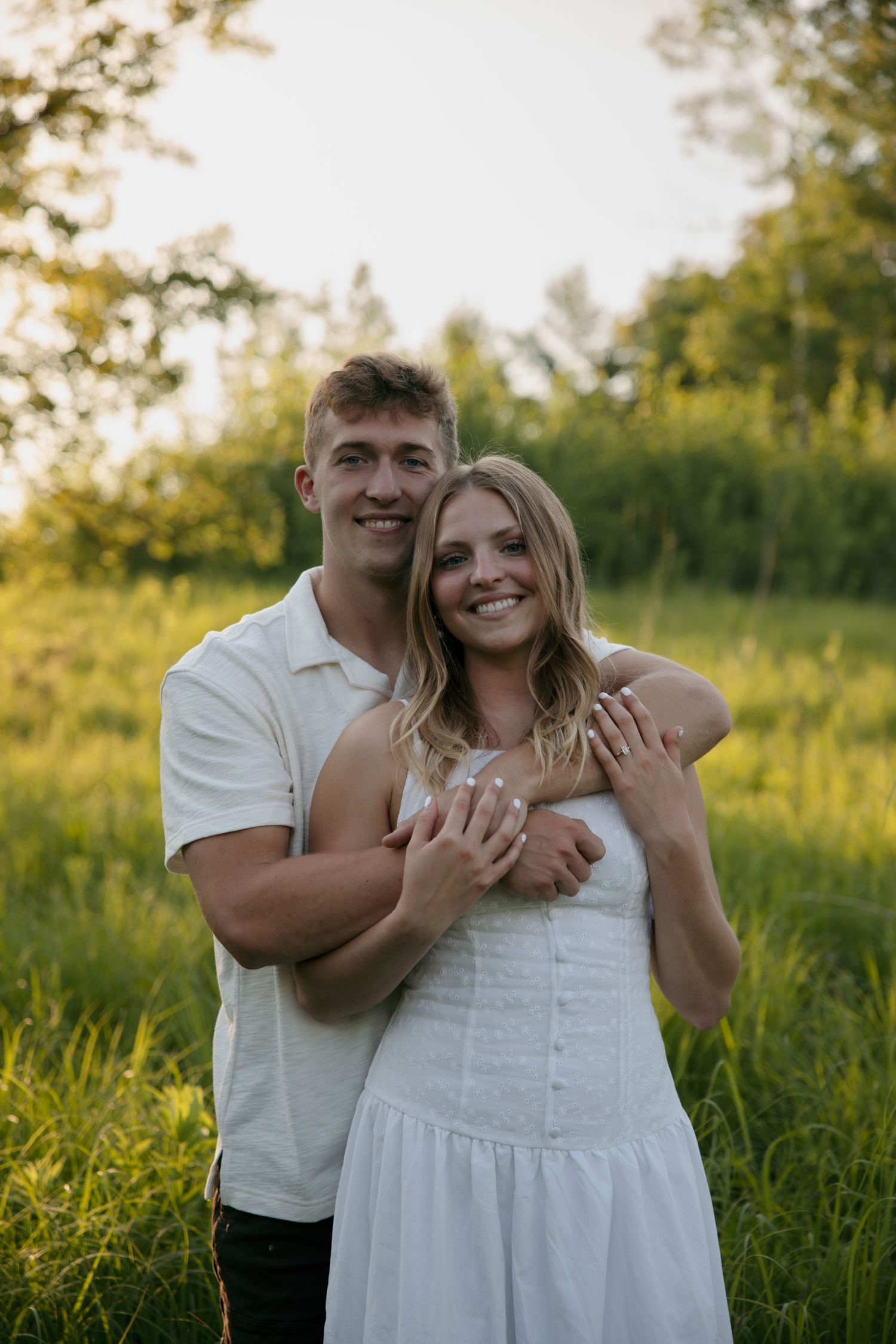 Couple embracing in a sunny green meadow, man in white shirt hugging woman in white dress, smiling at camera.
