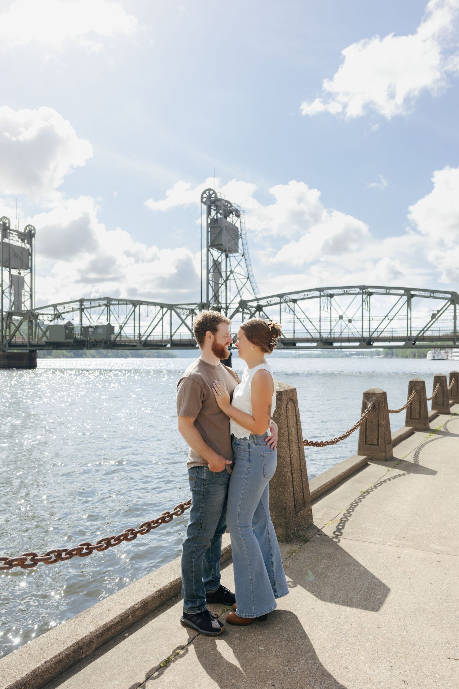 Couple embracing by a waterfront with a historic lift bridge backdrop on a sunny day.