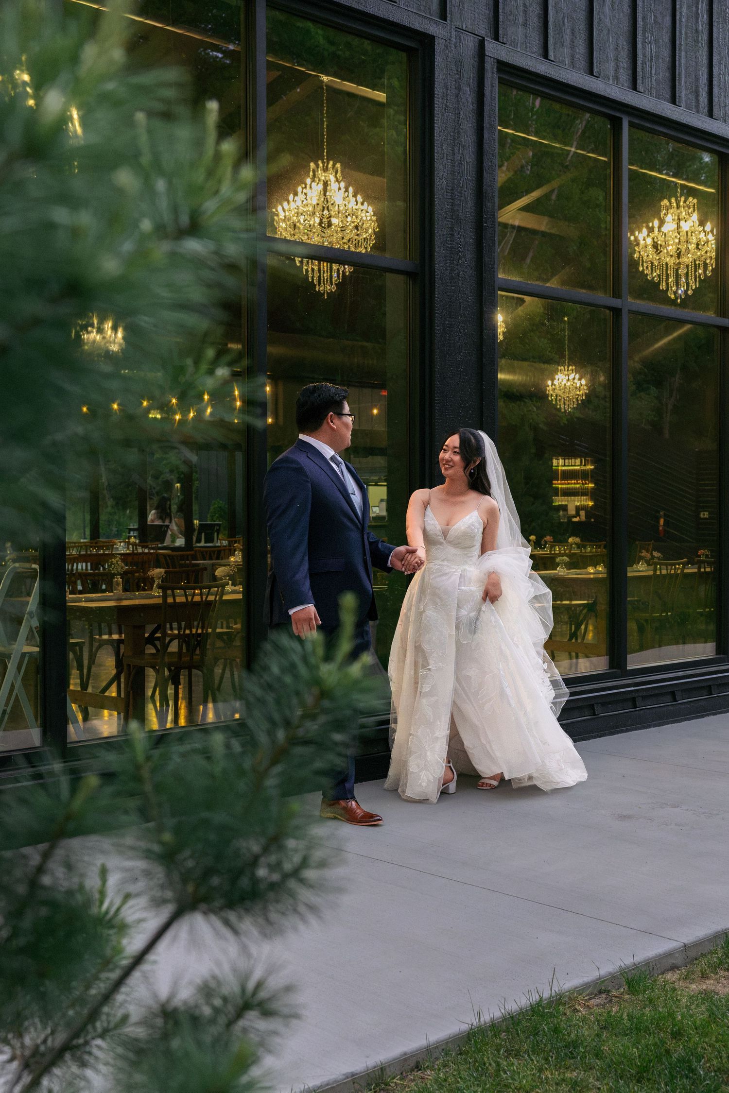 Bride and groom walking hand in hand outside elegant venue with glowing chandeliers visible through windows.