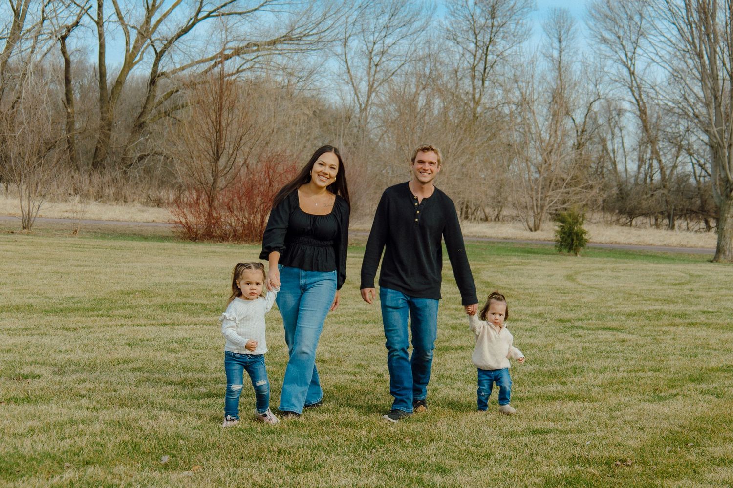Family of four walking in a green park, parents holding toddlers hands, bare trees in background.