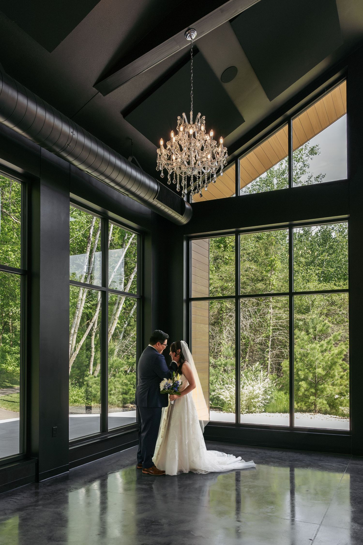 Bride and groom share a romantic moment under a crystal chandelier in a modern venue with floor-to-ceiling windows.