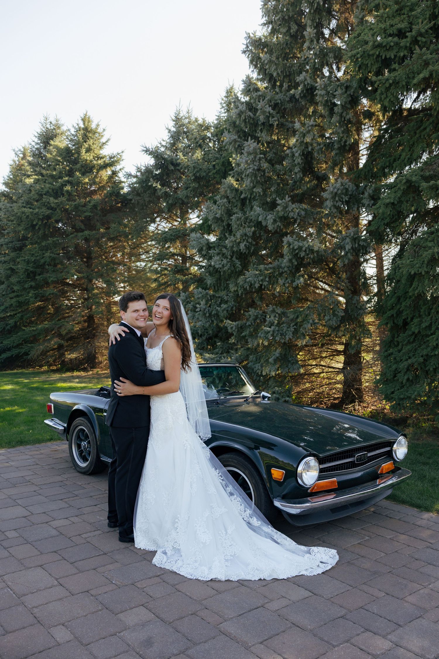 Bride and groom embracing in front of a vintage green car surrounded by tall evergreen trees on their wedding day.