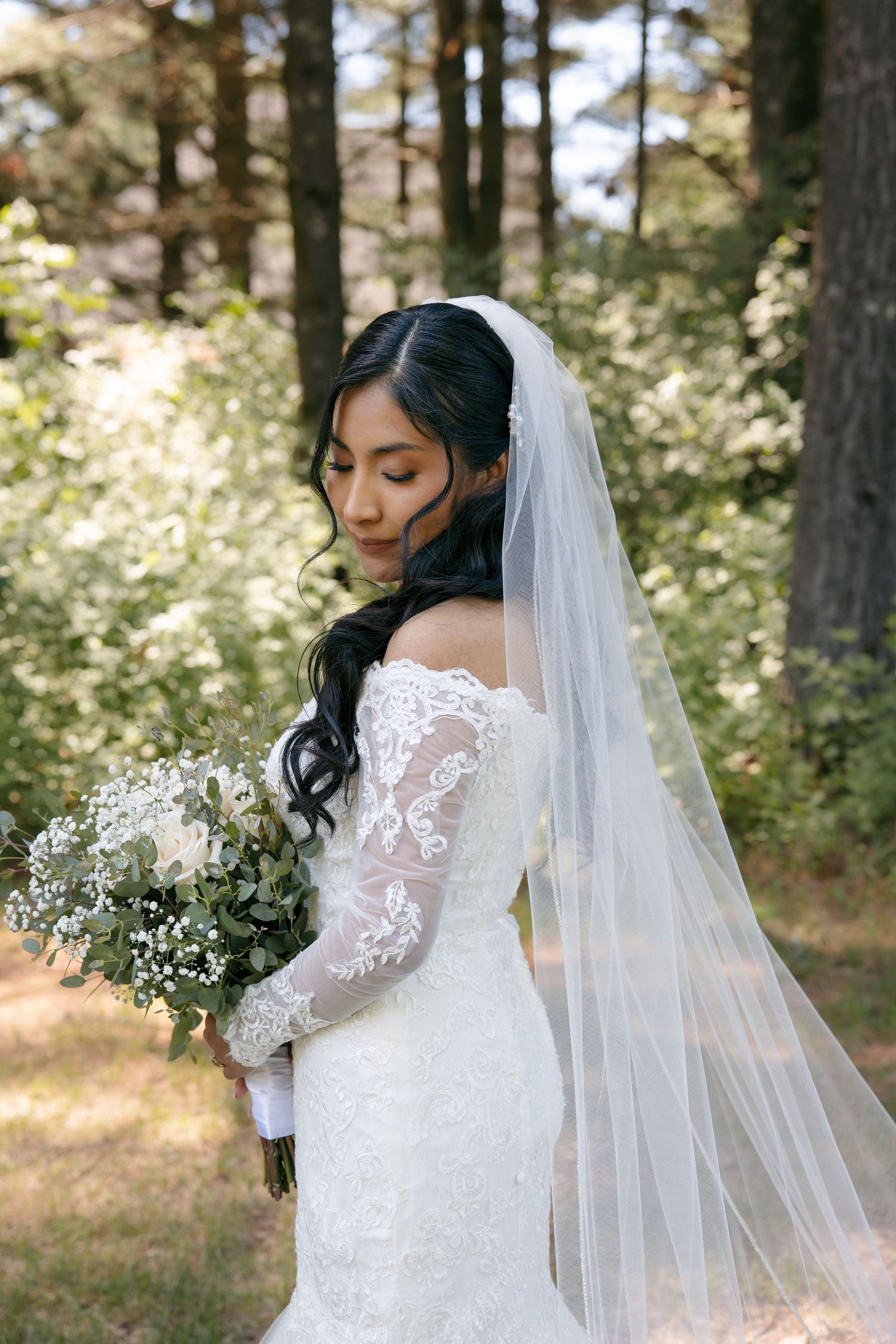 Bride in lace off-shoulder gown with veil holds white floral bouquet in sunlit forest setting.
