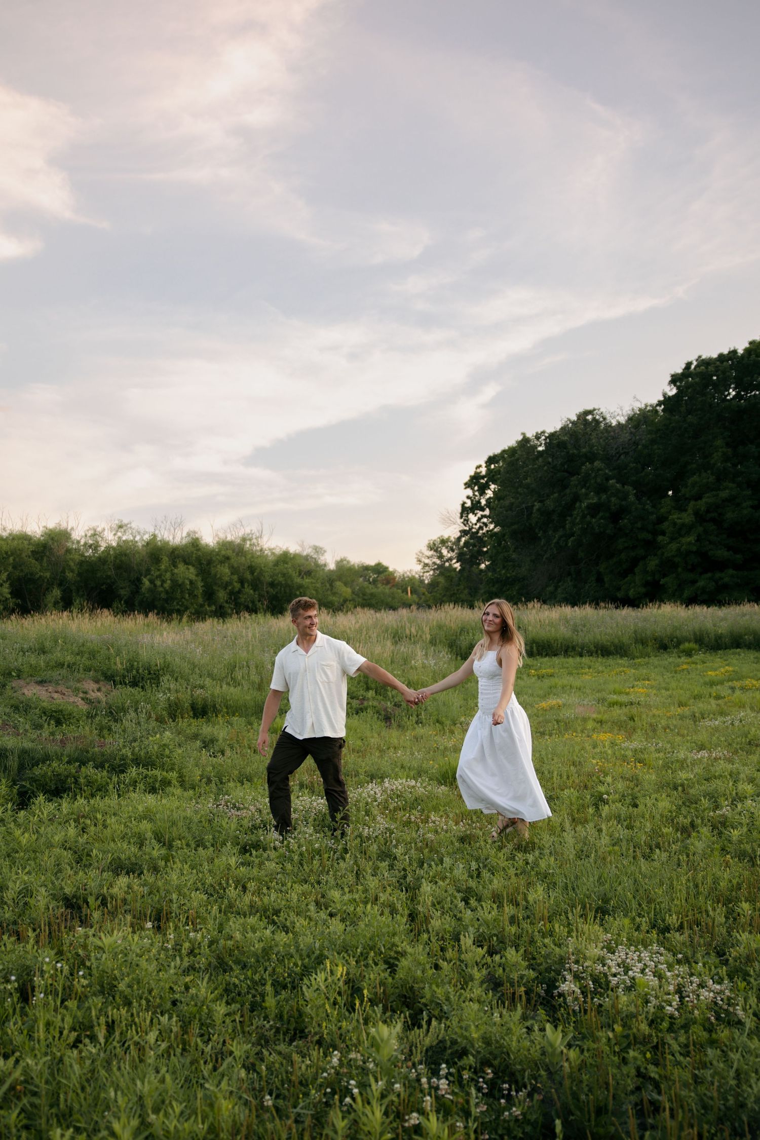 A couple holds hands walking through a lush green meadow at sunset, woman in white dress, man in white shirt.